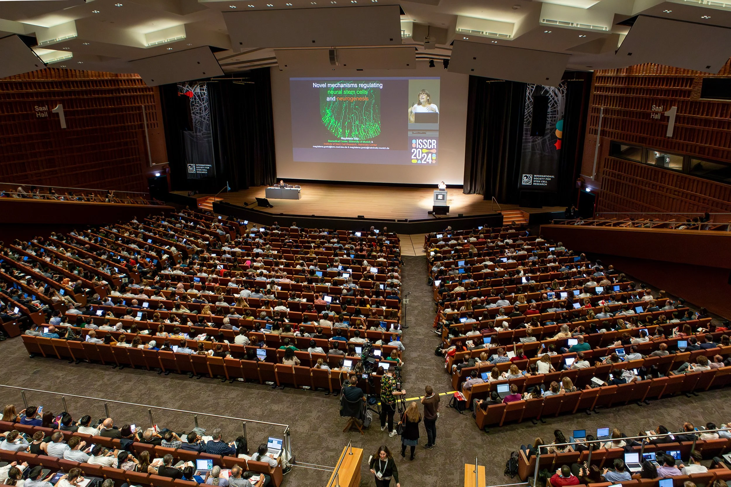 A large conference hall filled with hundreds of attendees watching a presentation on stage, with a woman speaking at a podium and a large screen displaying a scientific slide about neural stem cells and neurogenesis.