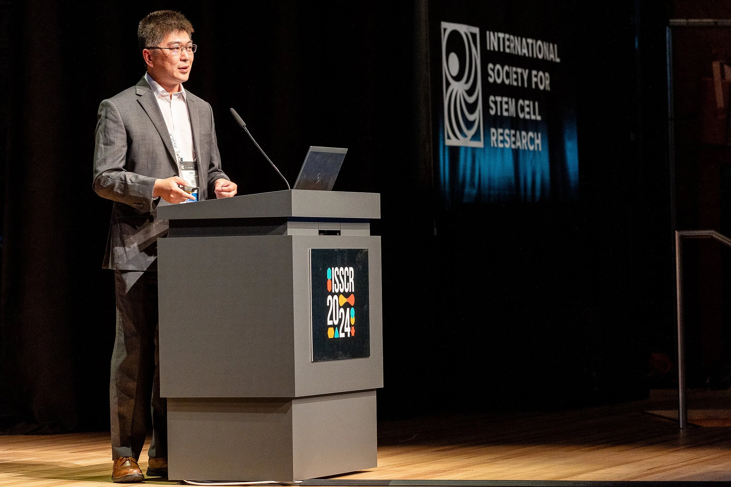 Man in a gray suit giving a presentation at a conference, standing behind a podium with a laptop at the ISSCR 2024 conference, with the International Society for Stem Cell Research logo on the screen in the background.
