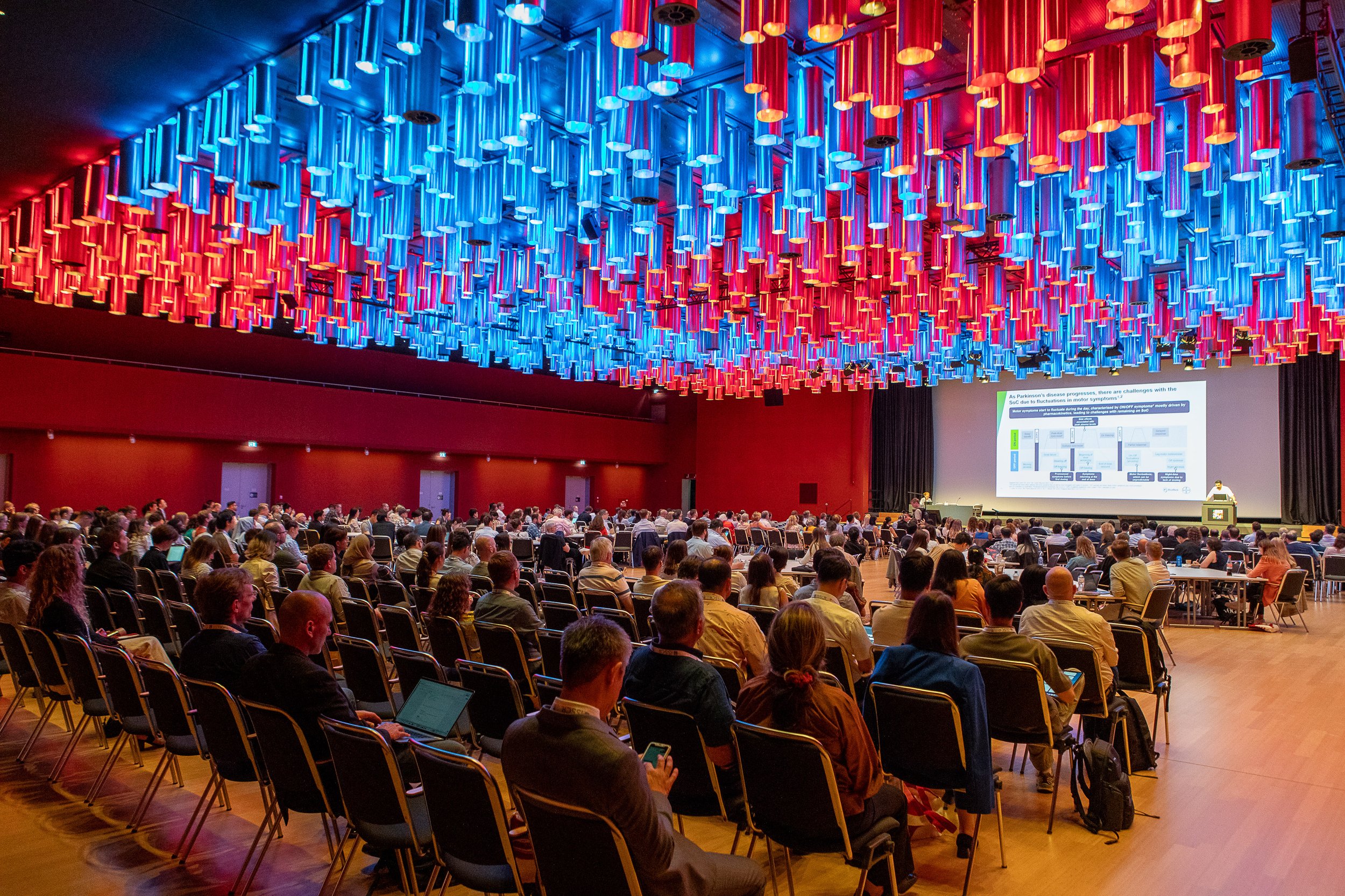 A large conference hall filled with seated attendees watching a presentation on stage. The ceiling features an elaborate installation of red and blue cylindrical lights.