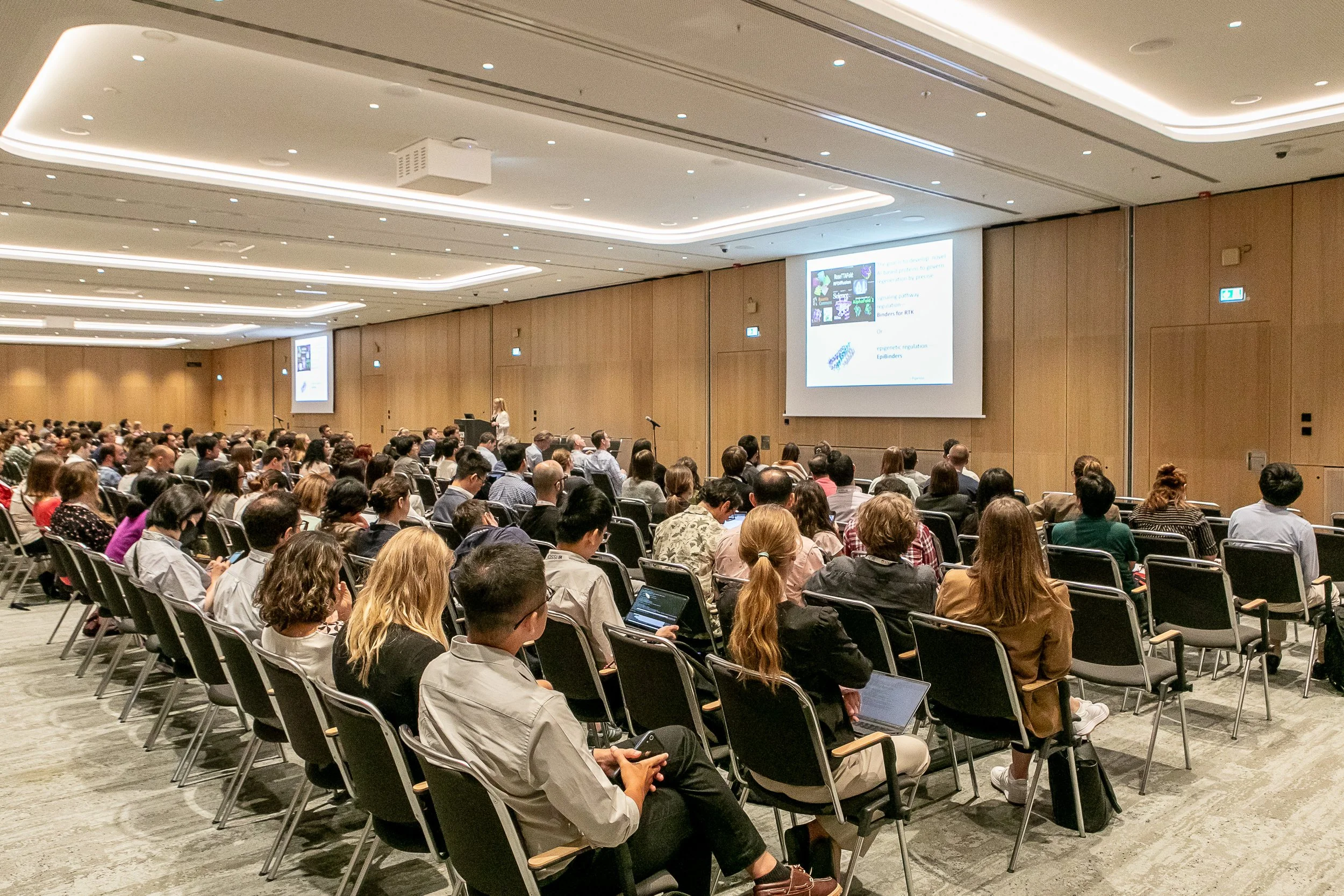 A large conference room filled with seated attendees watching a presentation on a large screen at the front.