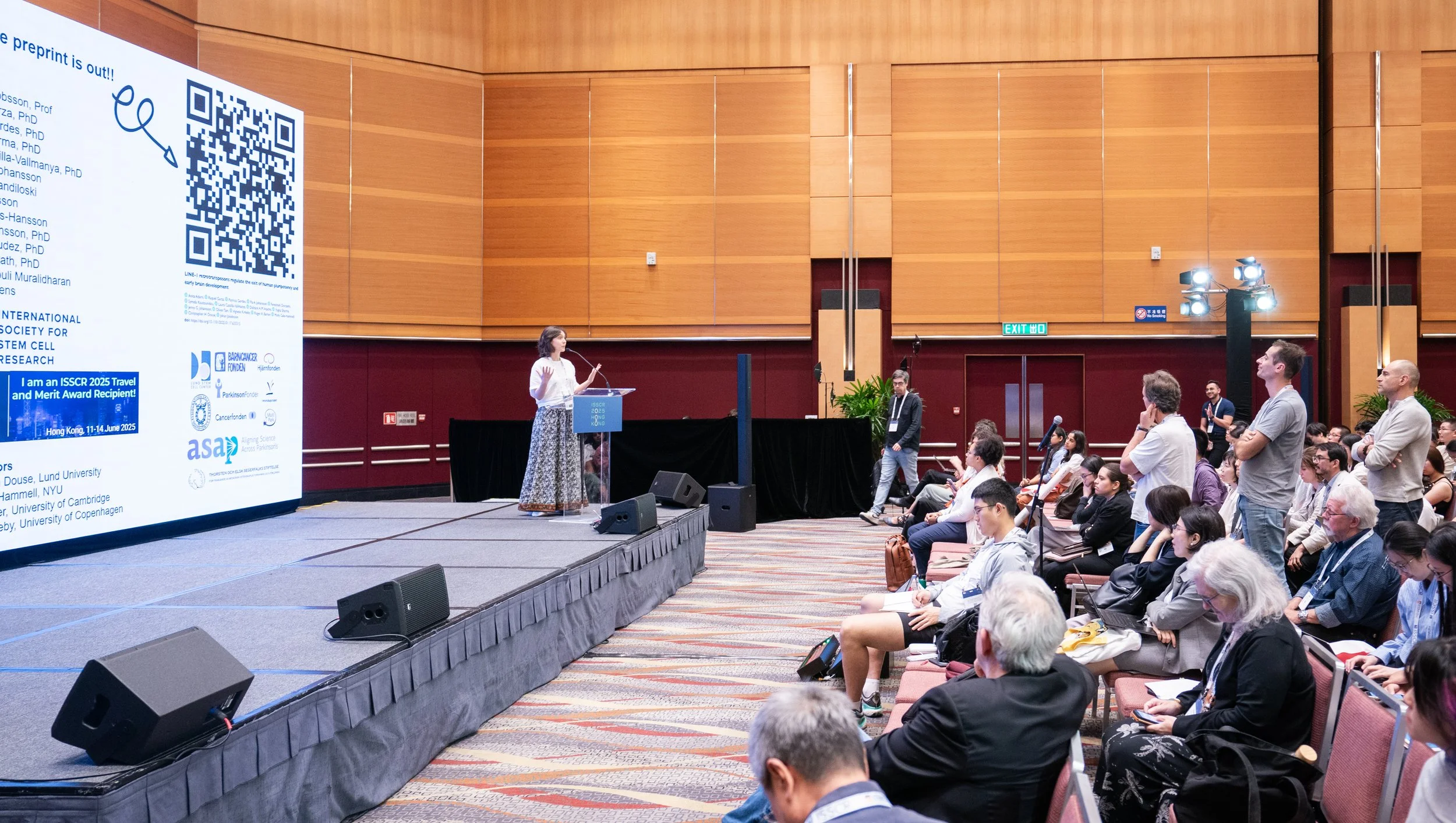 A woman stands at a podium on a stage, presenting to an audience in a conference room with wood-paneled walls. The large screen behind her displays a QR code and text about a scientific conference, including mentions of international awards and University affiliations. Audience members are seated, some taking notes, and some are standing or walking around.