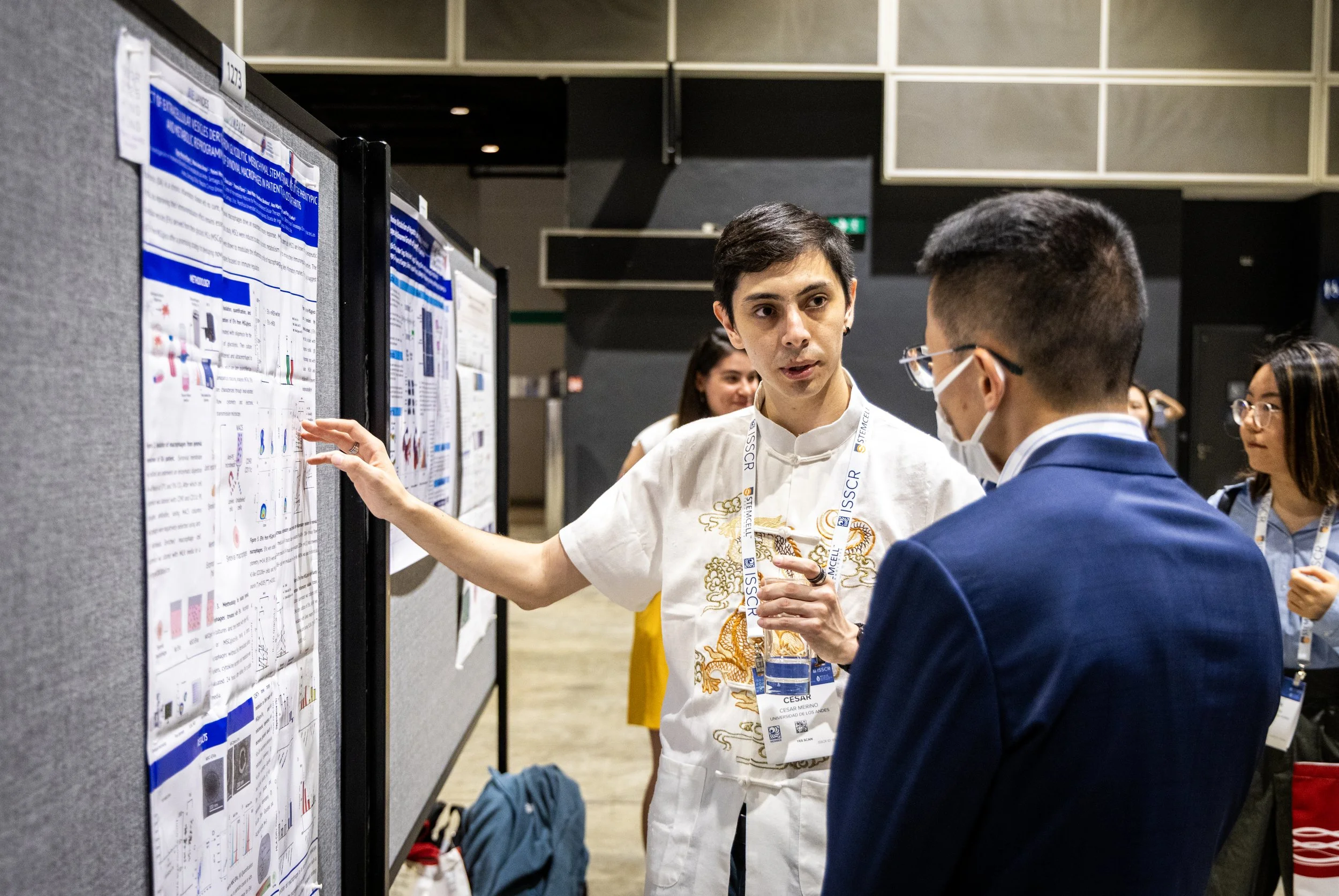 A man in traditional clothing is explaining scientific posters to another man wearing a mask at a conference