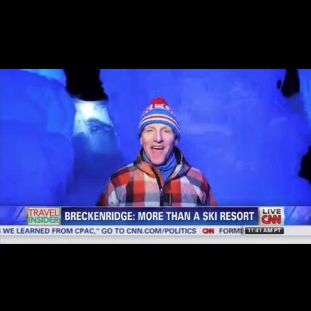 A journalist in a plaid jacket and winter hat smiling in front of an Ice Castle during a CNN broadcast about winter activities near Breckenridge ski resort.