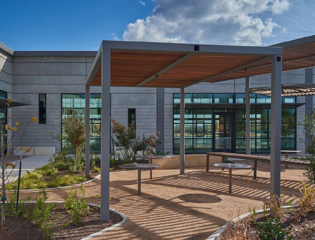 Outdoor seating area with a modern building in the background, a shaded pergola structure, and a landscaped garden with plants and bushes under a partly cloudy sky.