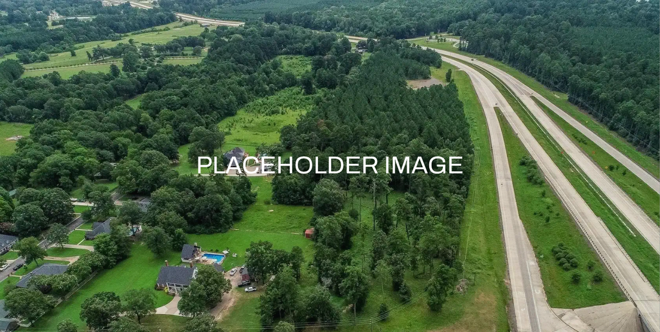 Aerial view of a residential neighborhood with houses, trees, and a swimming pool, adjacent to a highway surrounded by green forested areas.