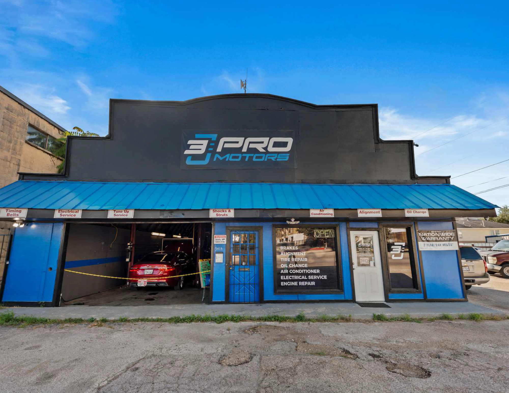 Front view of an auto repair shop named 3 PRO Motors with a red car inside, a blue exterior, and signs advertising services like brakes, alignment, tire repair, oil change, air conditioning, electrical service, and engine repair.