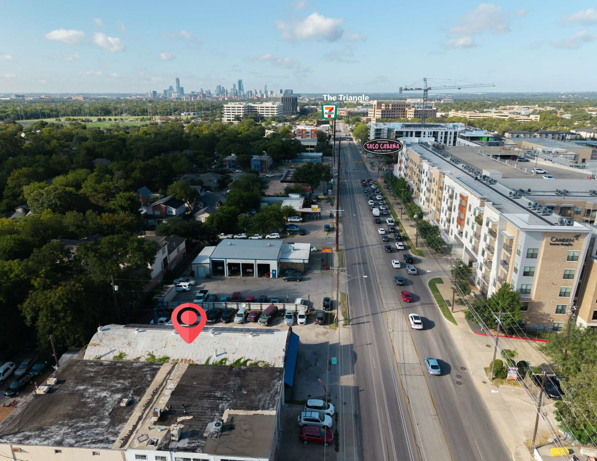 An aerial cityscape of a commercial and residential area with a busy street filled with cars, buildings including a CVS pharmacy, condos, and a construction crane in the background with the skyline of a city in the distance under a partly cloudy sky.
