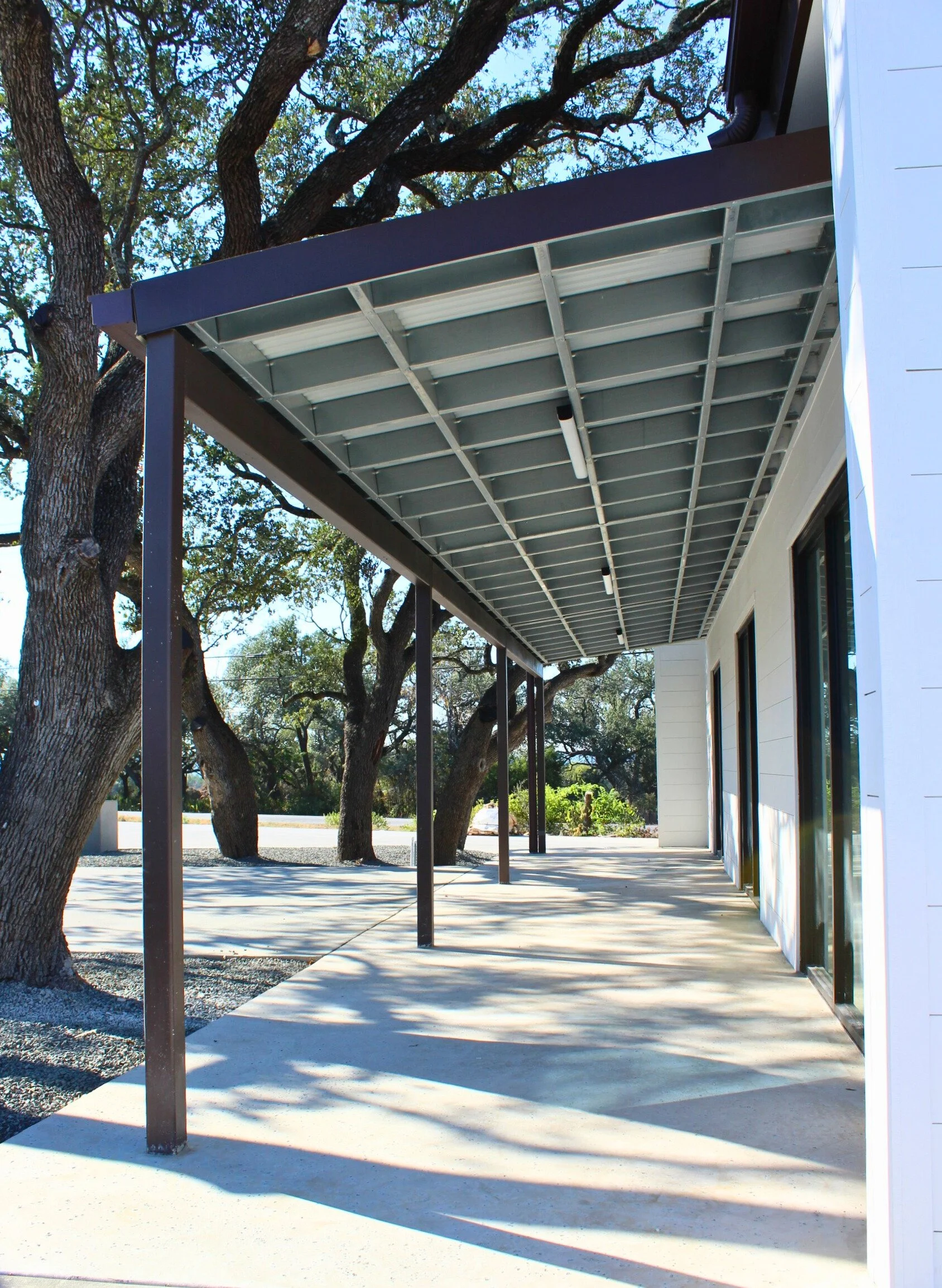 Empty building exterior with a covered patio, metal supports, and trees in the background under a clear sky.