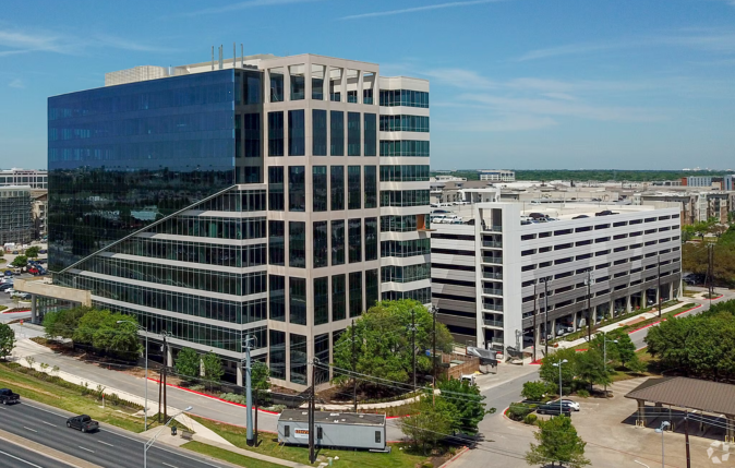 A modern glass office building with a distinctive sloped design and several floors, surrounded by trees and parking lots under a clear blue sky.