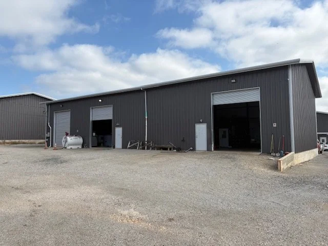 Exterior of a large, dark gray metal industrial building with three garage doors, on a gravel lot under a partly cloudy sky.