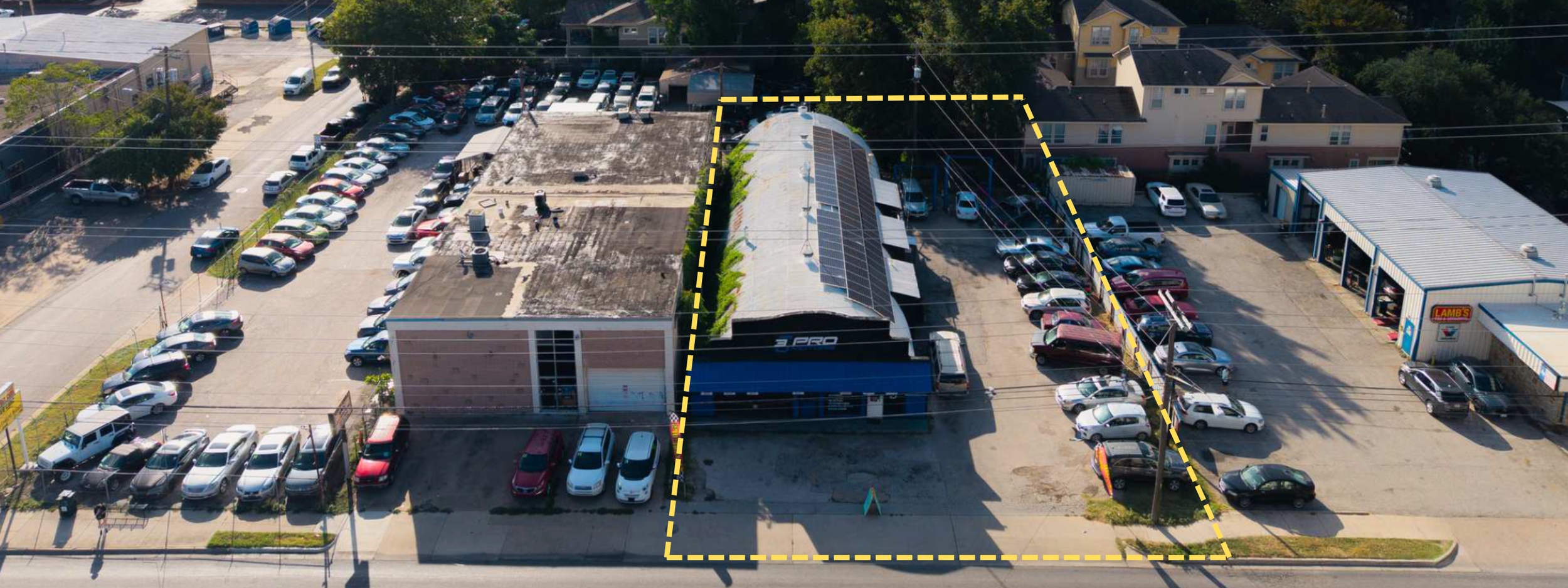 Aerial view of a commercial parking lot with multiple rows of cars, adjacent to a building with a solar panel roof, and an area outlined with dashed yellow lines.