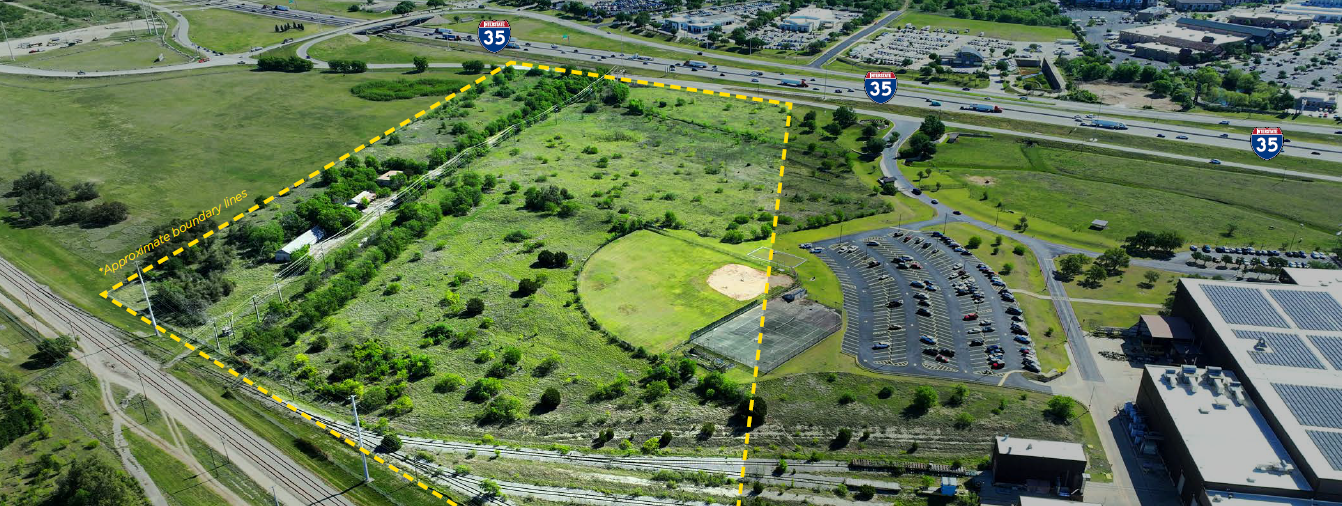 Aerial view of a large, open, green grassy area with trees bordered by a highway and a parking lot with multiple cars. Approximate boundary lines are marked with yellow dashed lines.