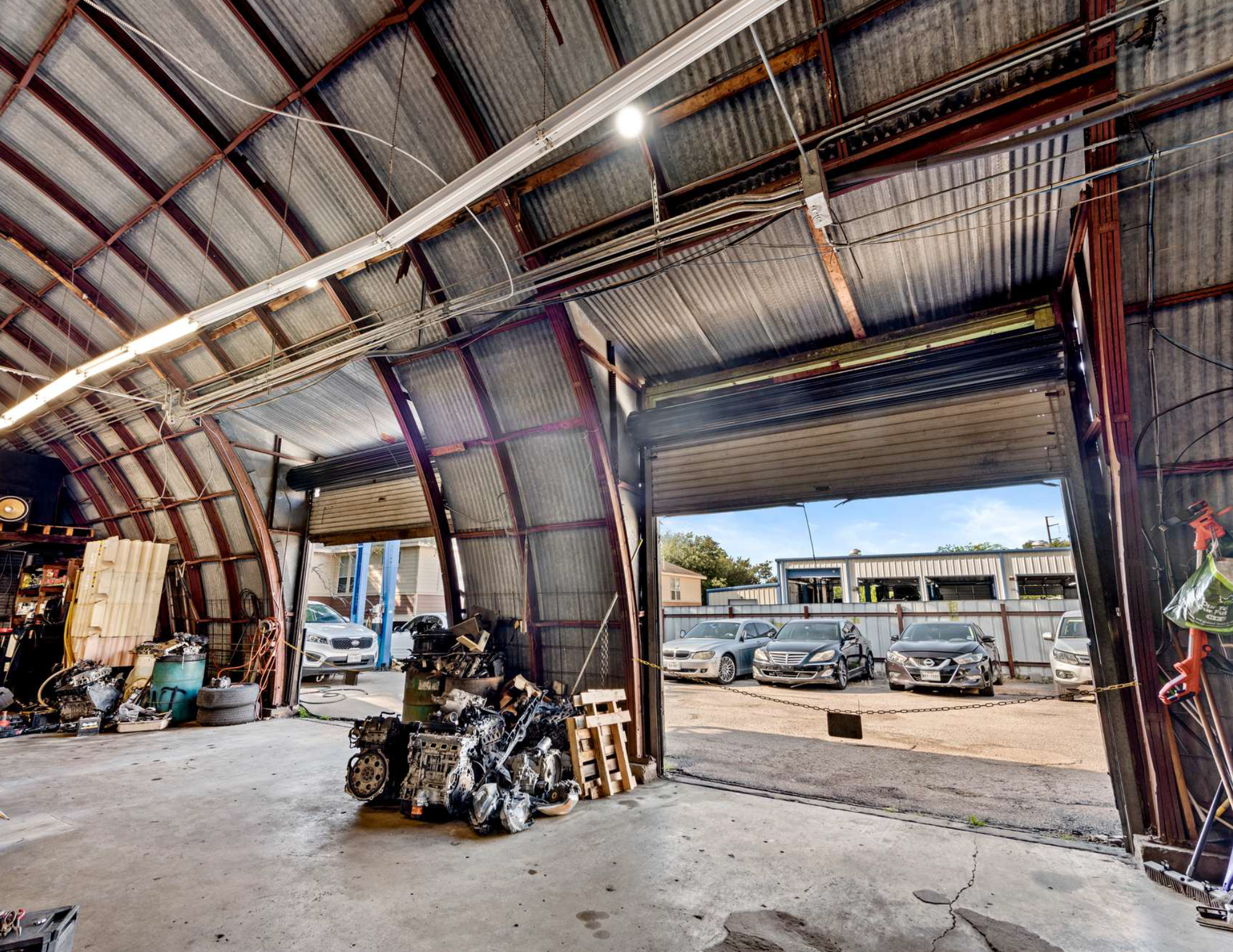 View inside a metal auto repair shop with a partially open garage door revealing parked cars outside, engine parts on the floor, and tools and equipment along the walls.