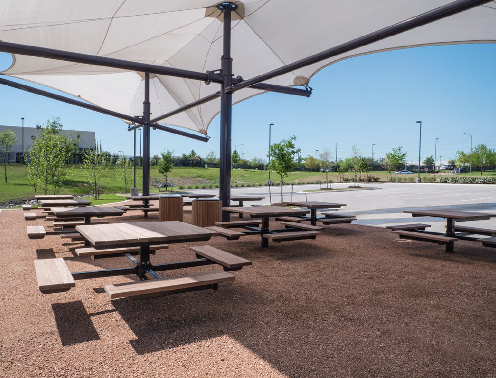 Empty outdoor picnic area with wooden tables and benches shaded by large white umbrellas, surrounded by green grass, small trees, and a parking lot in the background.