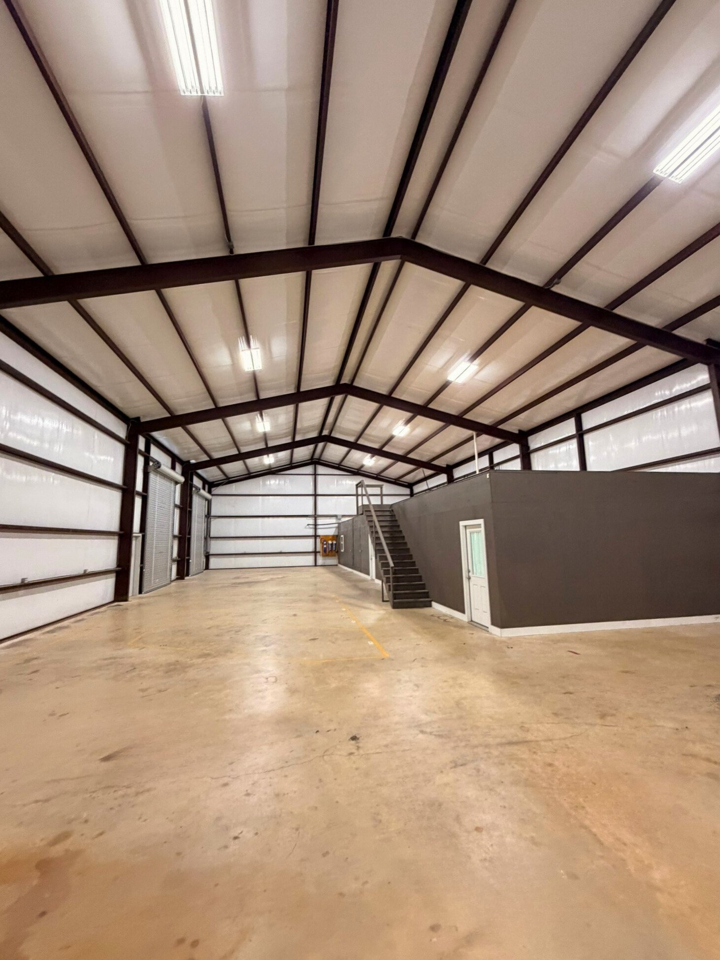 Large empty warehouse with metal walls and ceiling, concrete floor, and a small structure with stairs and a door on the right side.