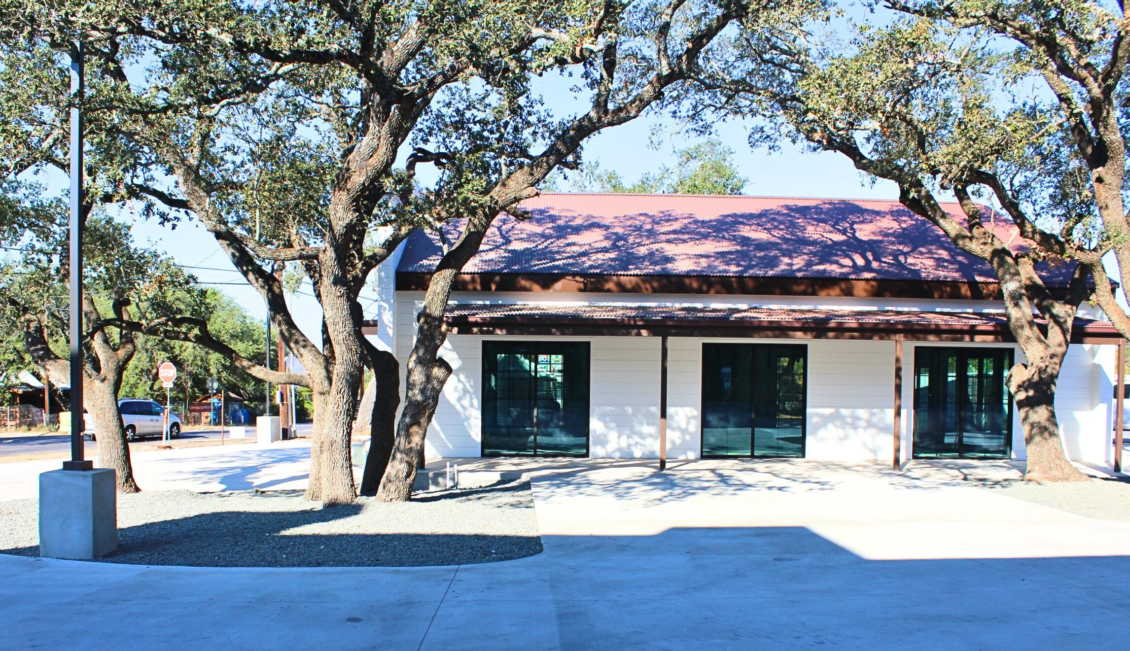 A building surrounded by several large trees casting shadows on the ground. The building has large glass doors and a sloped metal roof. The scene appears to be in a sunny area with a sidewalk and street nearby.