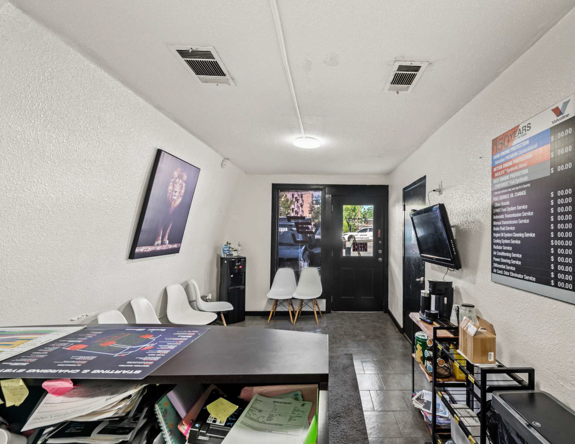Interior of a small waiting area with white chairs, a black door with a window, a wall-mounted TV, a framed artwork, a desk cluttered with papers, a table with beverages, a black rack with snacks, and a bulletin board with service prices.