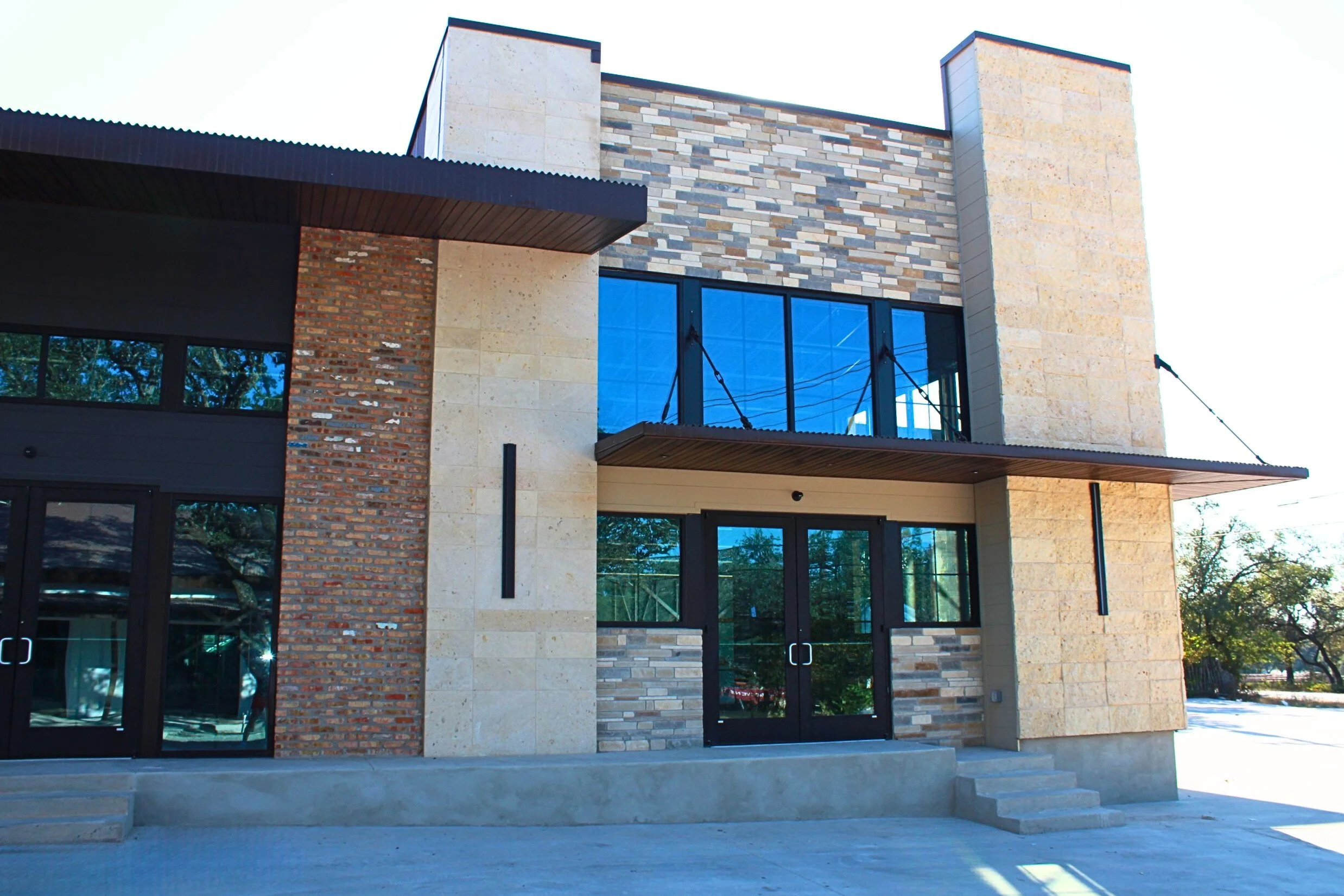 Modern building with a mix of brick, stone, and glass exterior, featuring large windows, glass doors, and a small outdoor staircase.