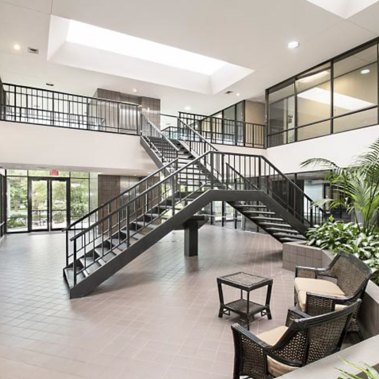 Indoor lobby area with modern black staircase, glass railings, large windows, and seating area with cushioned chairs and potted plants.