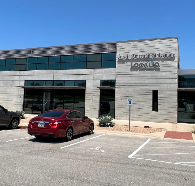 The exterior of a modern building with a sign that reads 'Austin American-Statesman' and 'LOCAL IQ', with parking spaces in front, including a red car and a blue handicap parking sign.