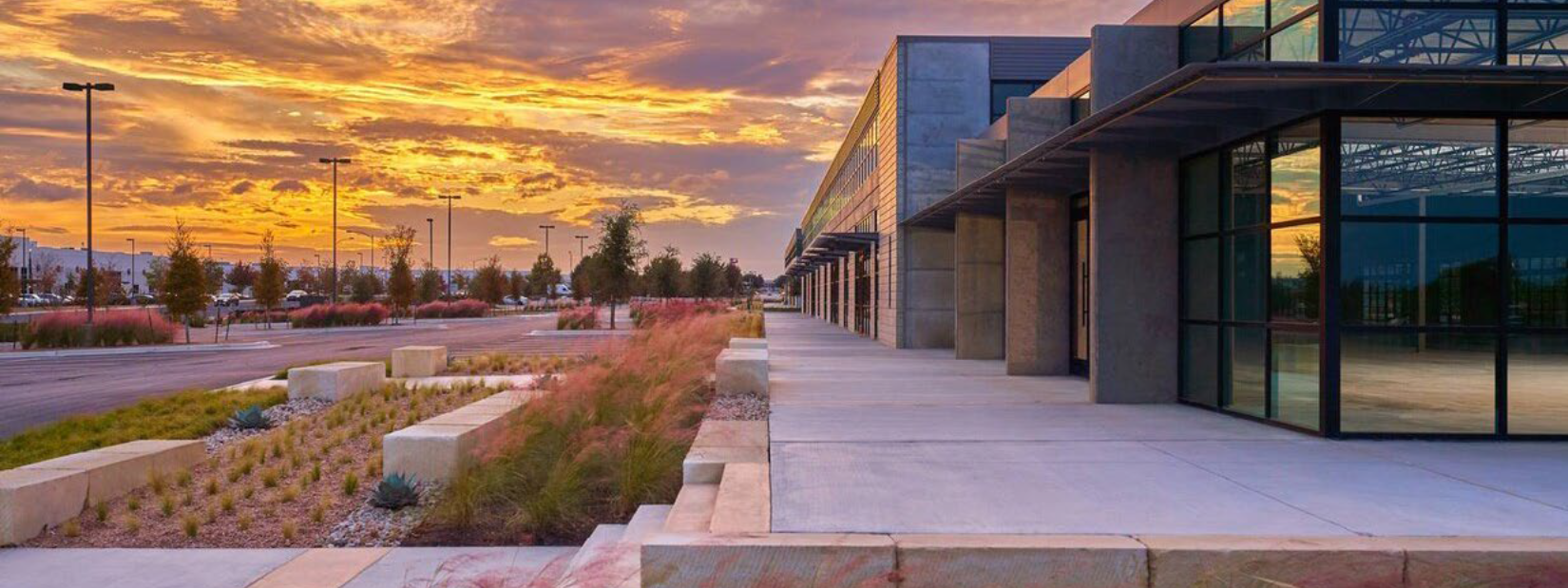 Modern commercial building with glass windows, concrete sidewalk, landscaped area with plants and rocks, and a parking lot, during sunset with an orange and purple sky.
