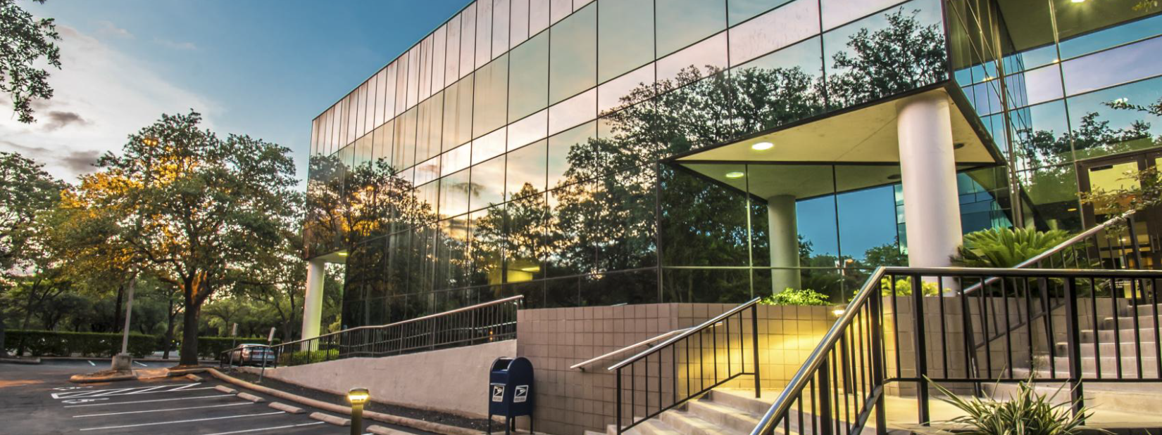 Photo of a modern office building with a glass facade reflecting trees and sky, located next to a parking lot and sidewalk with some plants and outdoor lighting.