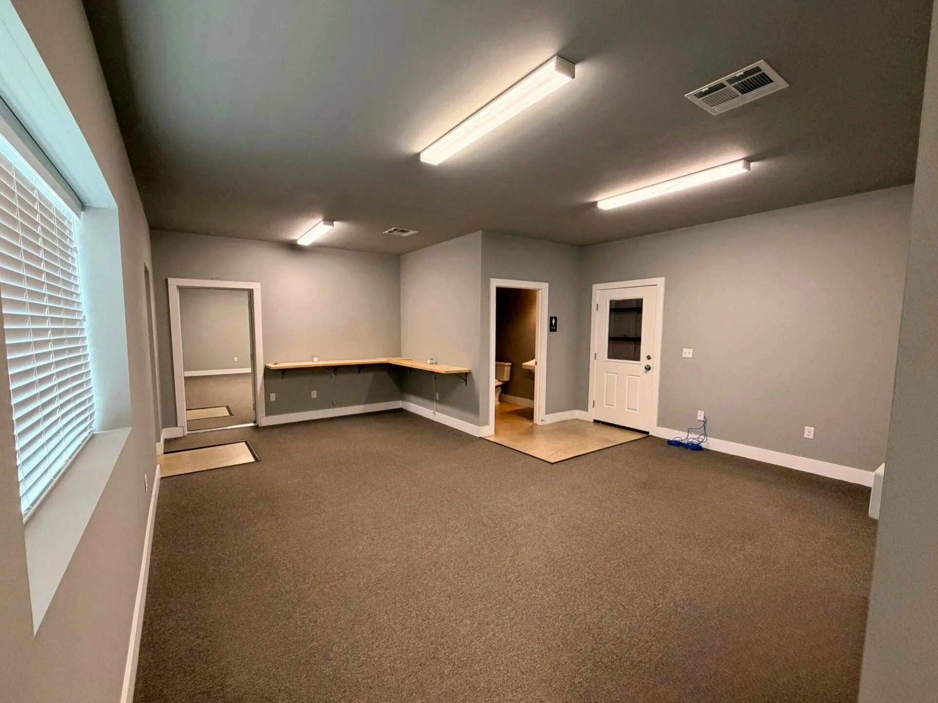 Empty office or meeting room with gray walls, brown carpet, window with blinds, fluorescent ceiling lights, an L-shaped built-in desk, and a small bathroom visible through a doorway.
