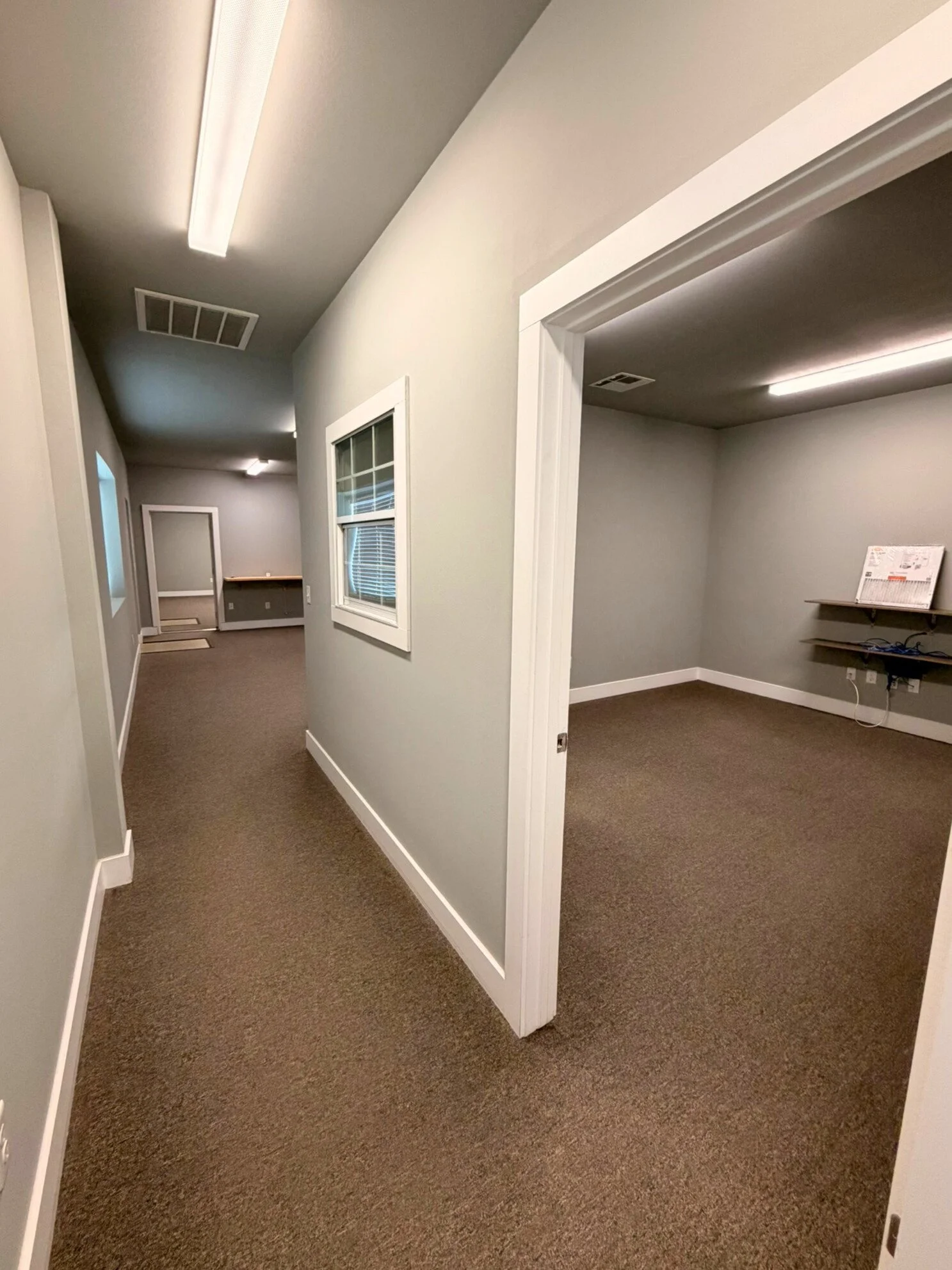 Interior view of an office hallway with beige walls, brown carpet, and white trim, showing windows and doorways leading to small rooms.