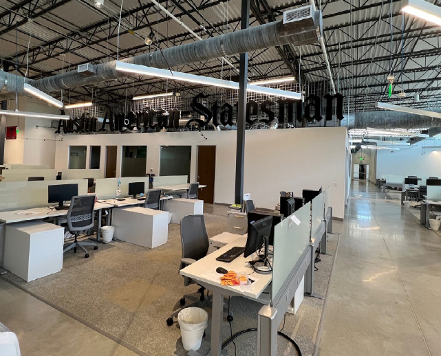 Open office space with multiple desks, computers, and office chairs, featuring exposed ductwork and a sign with black lettering on the wall reading "Salesian."