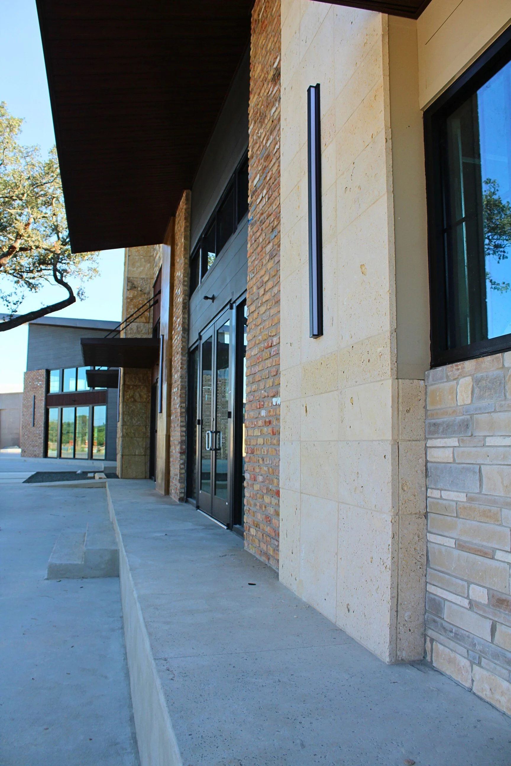 Modern commercial building with brick and stone exterior, black-framed glass doors, and tall wall-mounted light fixture, with a concrete walkway and trees in the background.