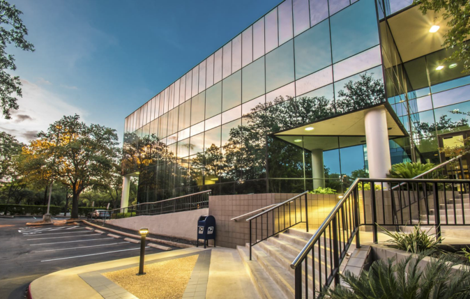 Modern glass office building with trees in the background, outdoor staircase and parking lot at dusk.
