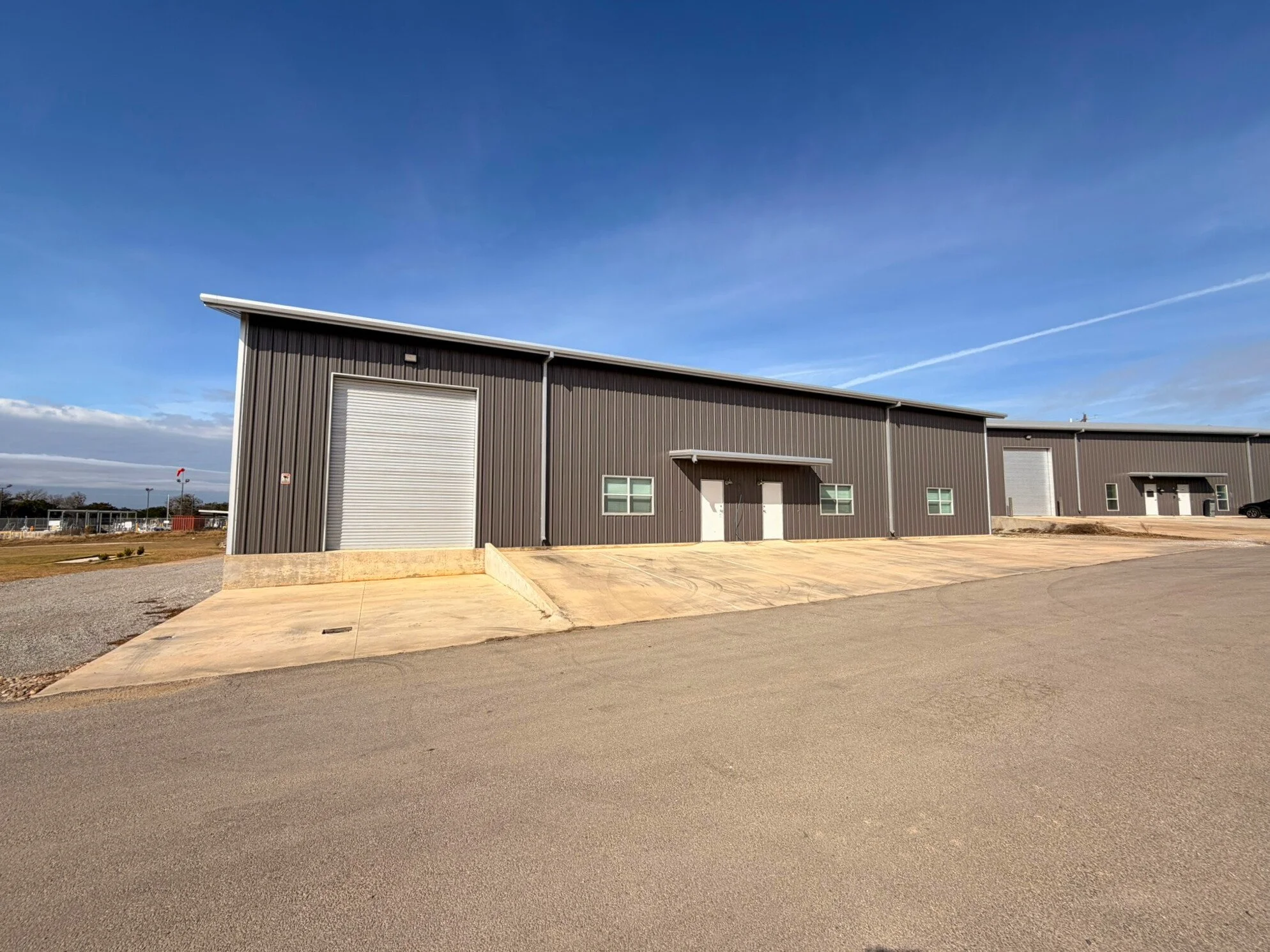 A large industrial warehouse building made of metal siding, featuring multiple large roll-up doors and small windows, situated on a paved lot under a blue sky with wispy clouds.