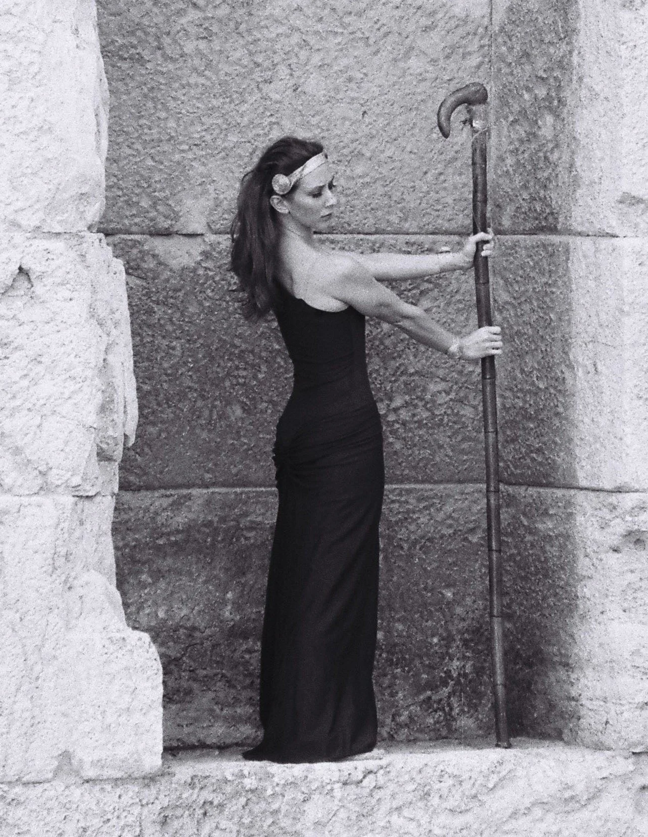 A woman in a long black dress holding a javelin against a rough stone wall.