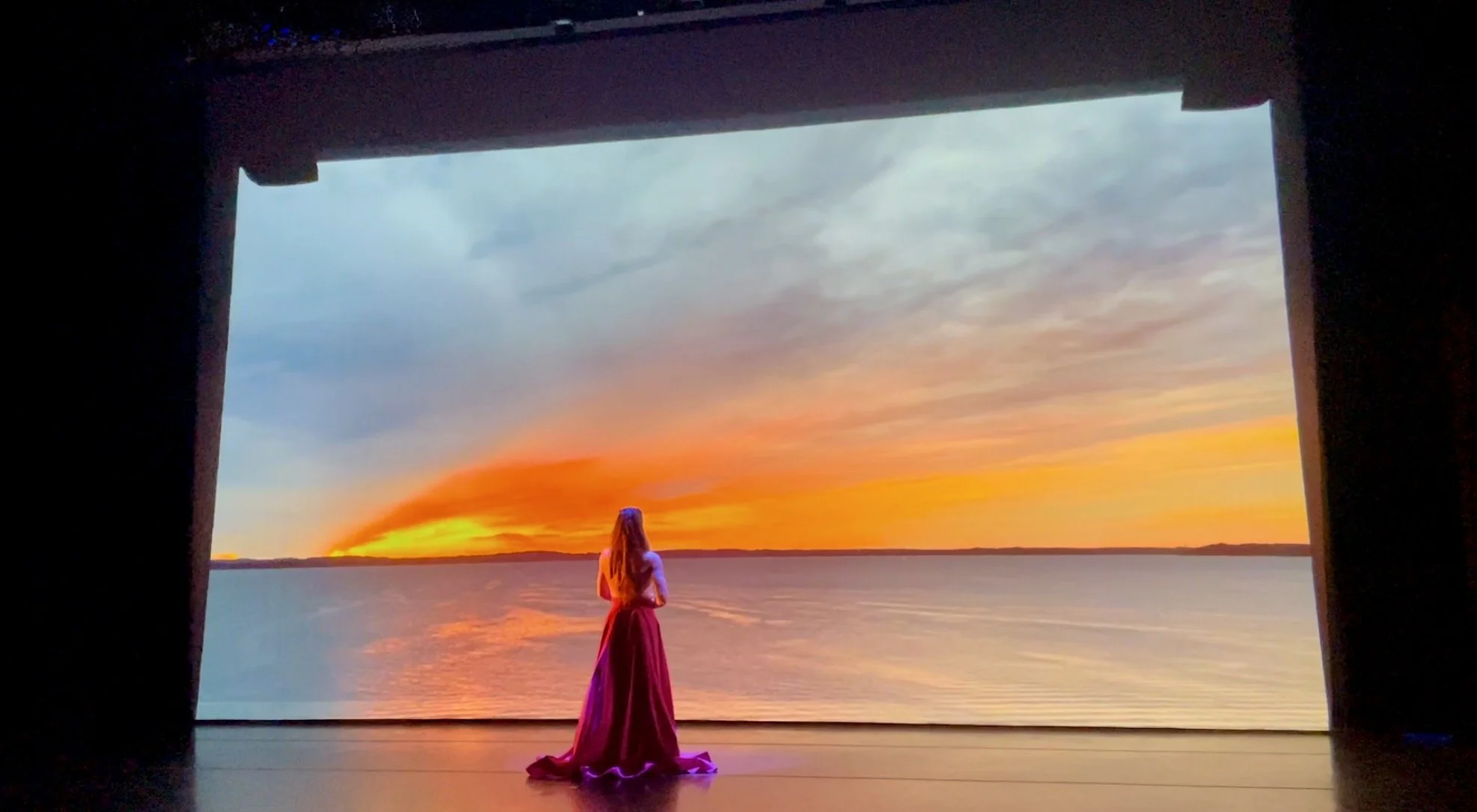 A woman in a pink dress standing on stage looking at a sunset over a body of water, framed by a stage opening.