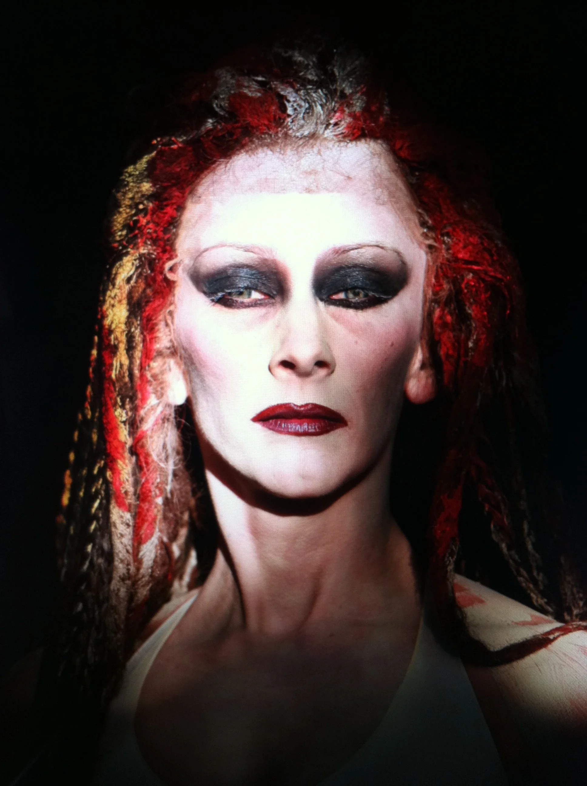 Close-up of a woman with pale skin, dark smoky eye makeup, red lipstick.