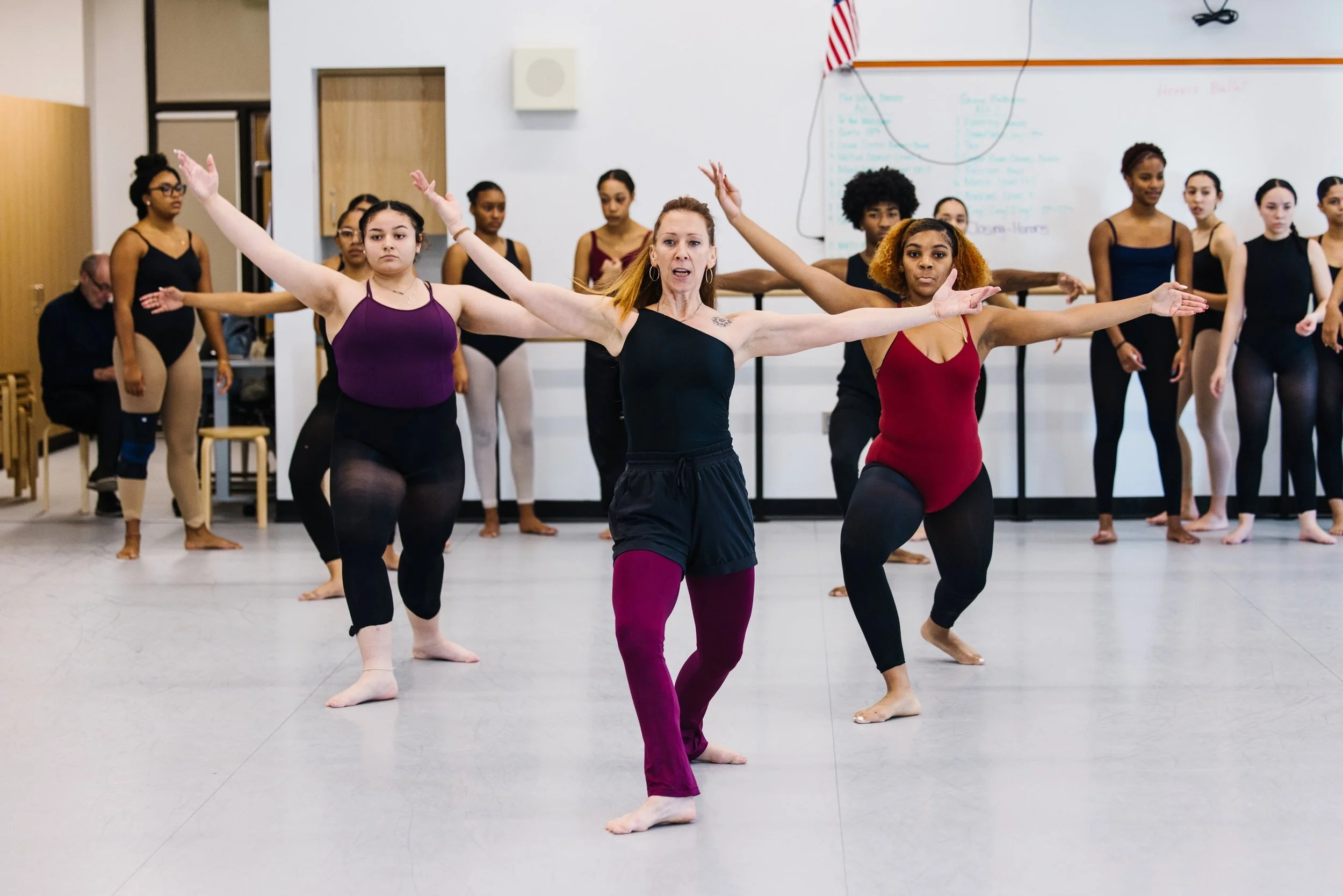 Dance instructor leading a group of women in a dance class, performing a move with arms extended to the sides in a studio with a whiteboard in the background.