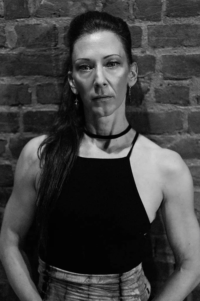 A woman with long dark hair standing against a brick wall, wearing a black sleeveless top and earrings.