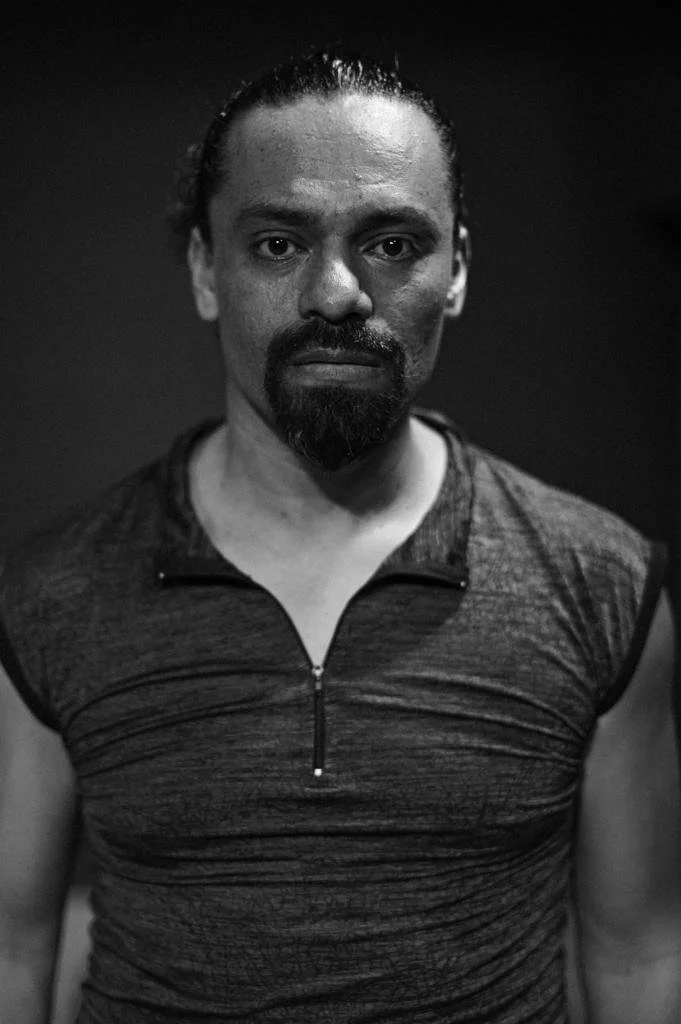 Black and white portrait of a man with a serious expression, dark hair pulled back, facial hair, wearing a textured athletic shirt with a zipper.