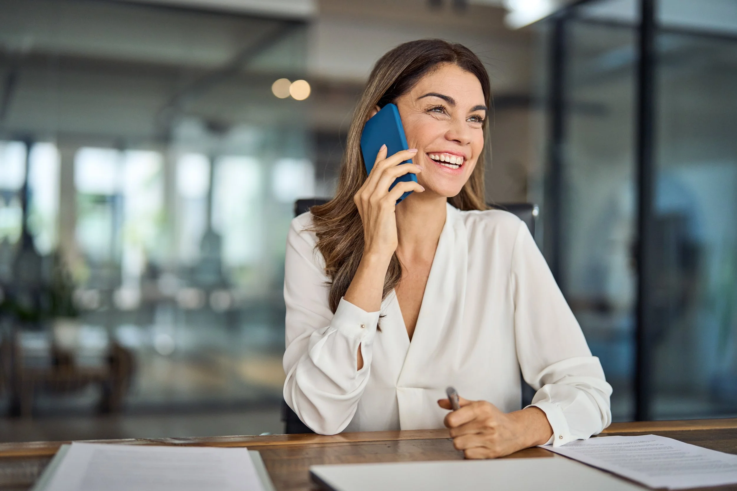 A woman with long brown hair is smiling and talking on a blue phone, sitting at a desk with papers in front of her in a modern office setting.