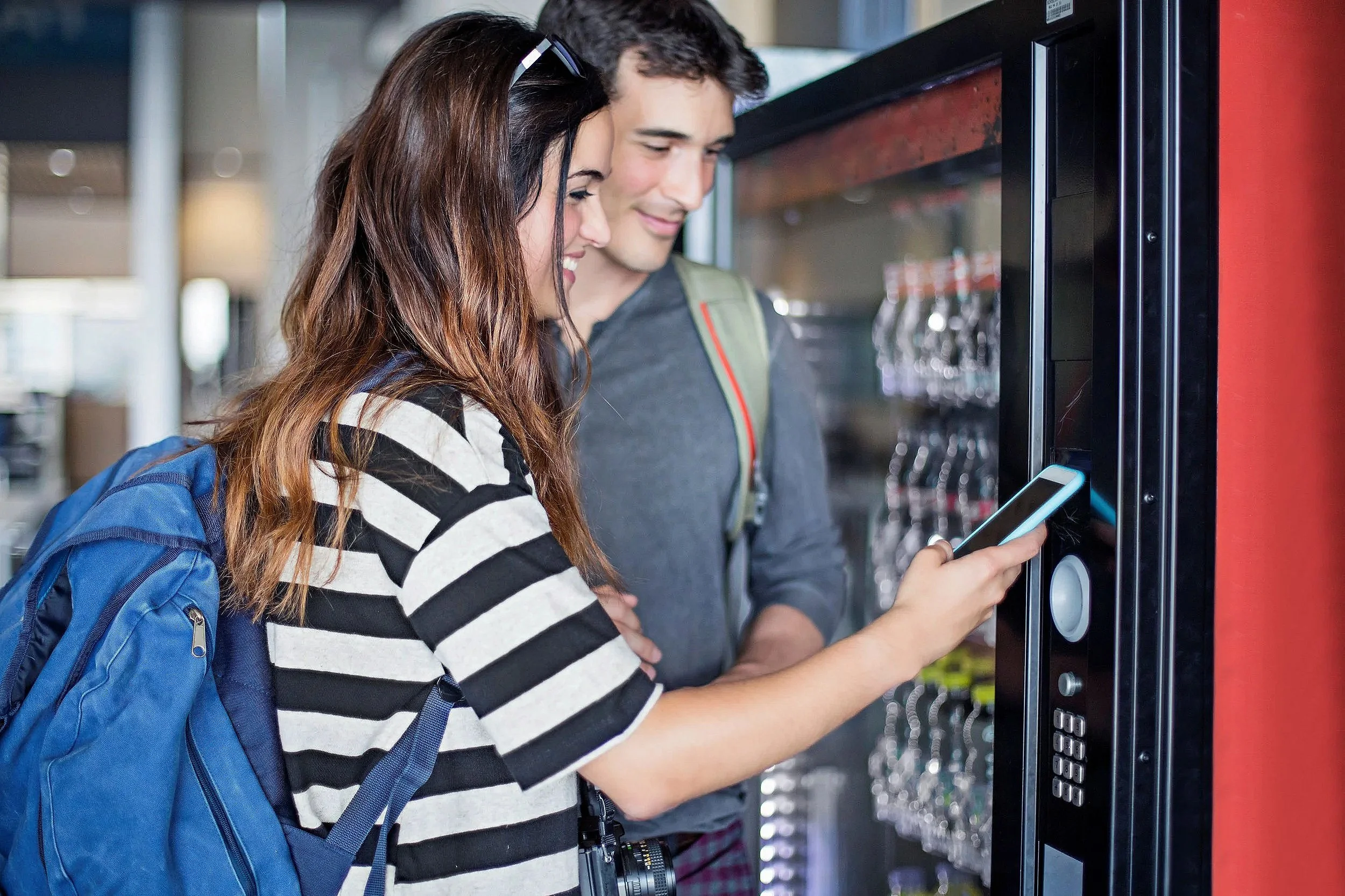 A young woman and a young man using a vending machine together in a modern indoor setting. The woman is holding a smartphone and smiling, while the man is looking at the vending machine, both appear happy and engaged.