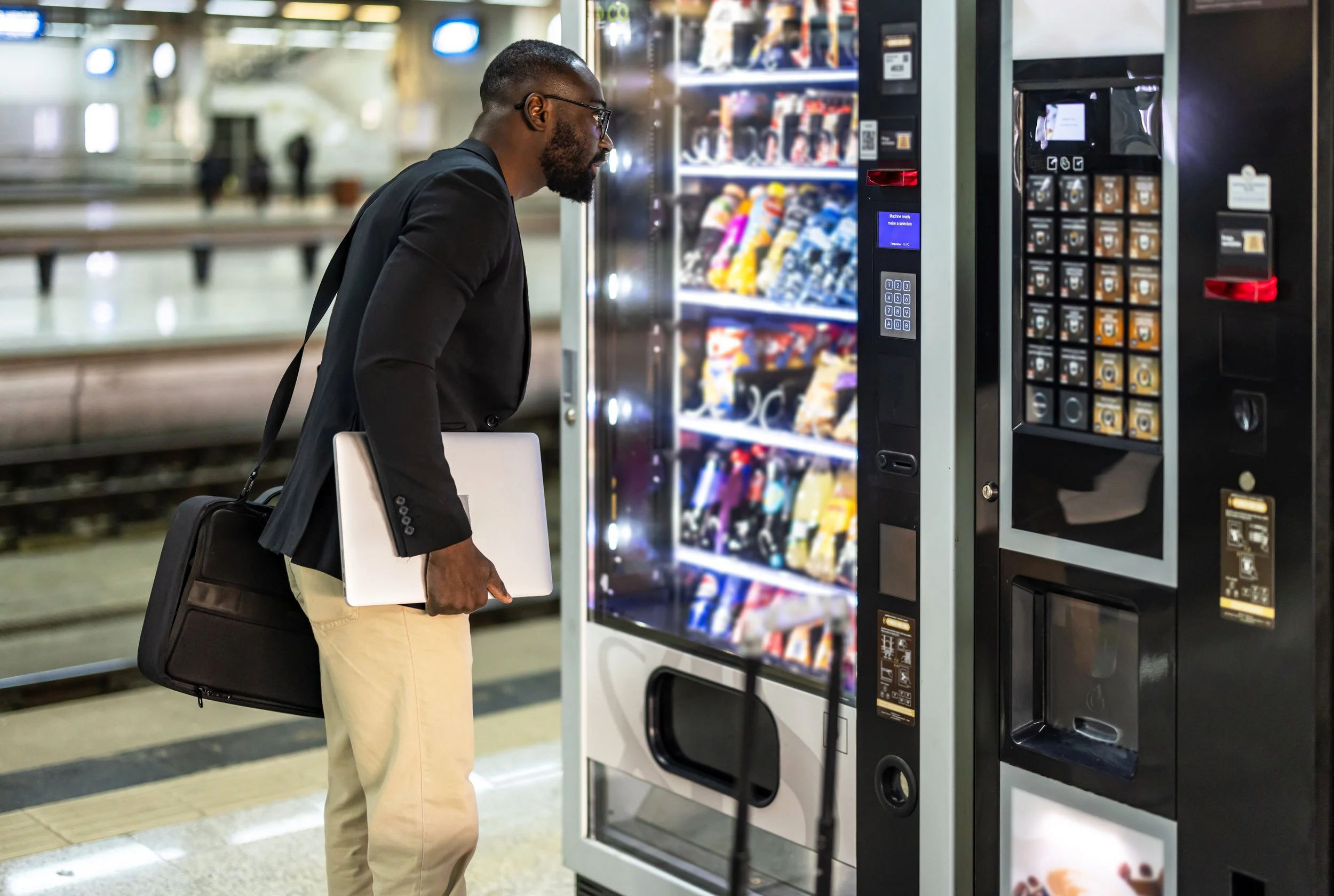 A man in beige pants and a black blazer with a laptop under his arm is using a vending machine at a train station.