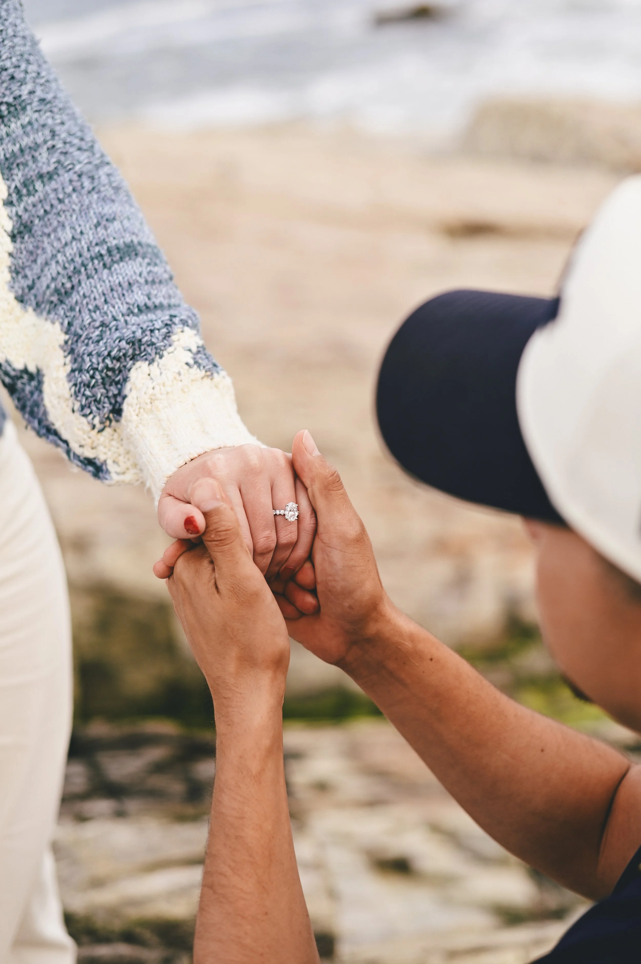 Detail shot of groom to be at the beach on one knee holding the hand of his future bride after placing the engagement ring on her finger.