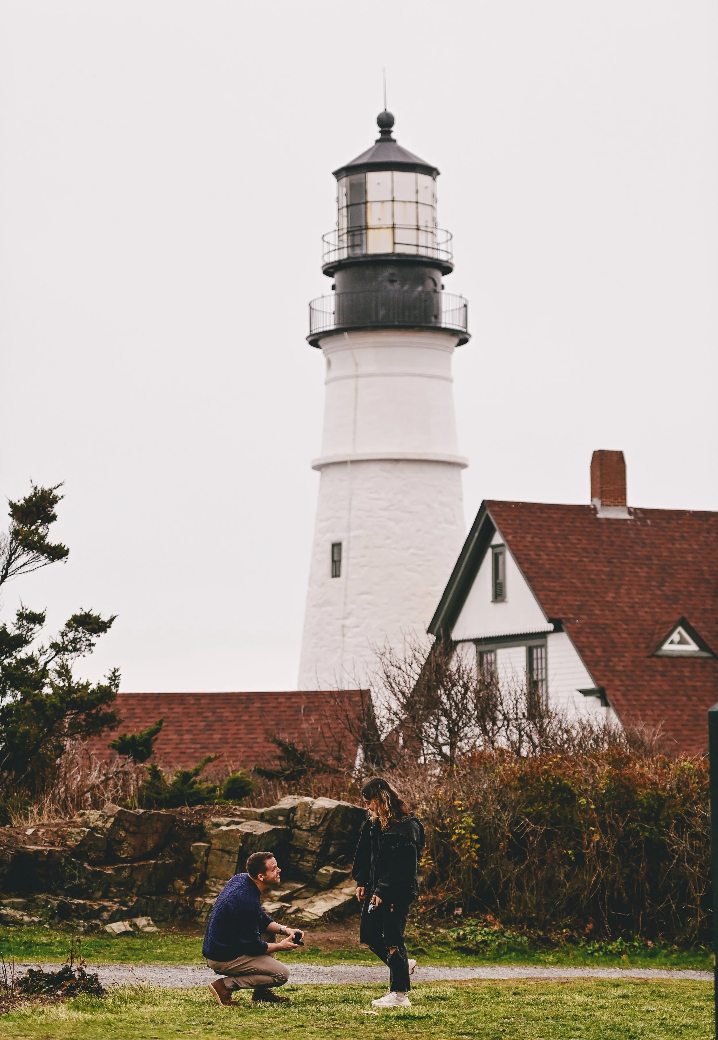 Future groom proposes on one knee in front of Portland Headlight lighthouse.
