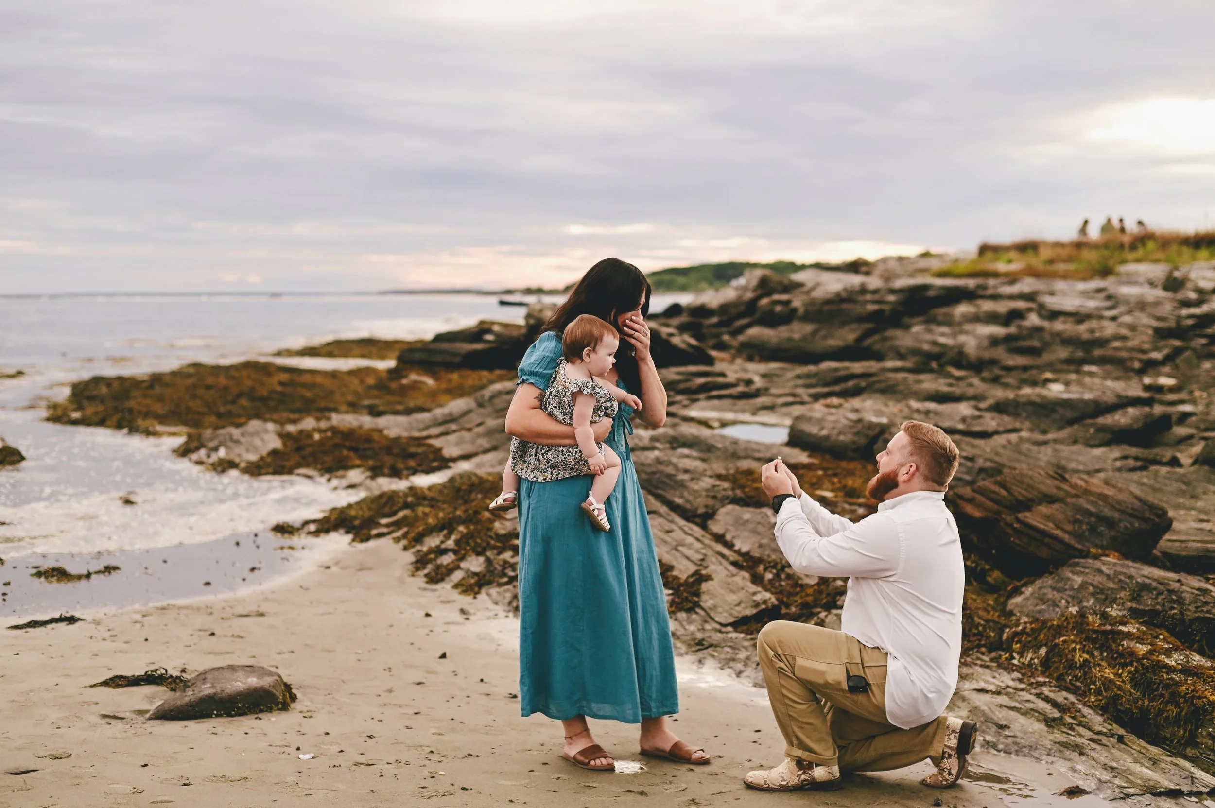 A coastal beach proposal with future bride surprised and holding baby while groom to be proposes on one knee.