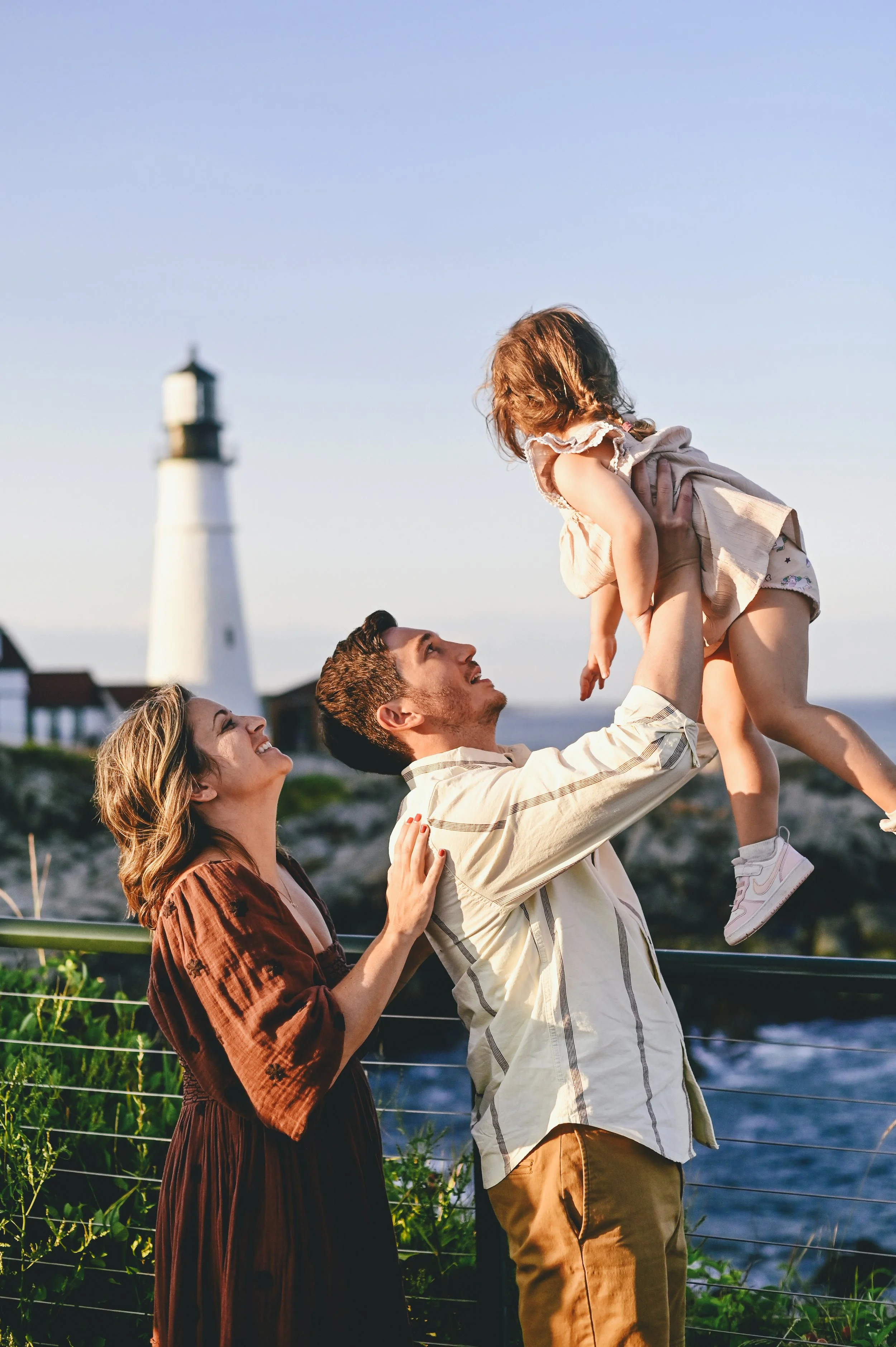 Family lifting their toddler in the air by Portland Head Light in Maine