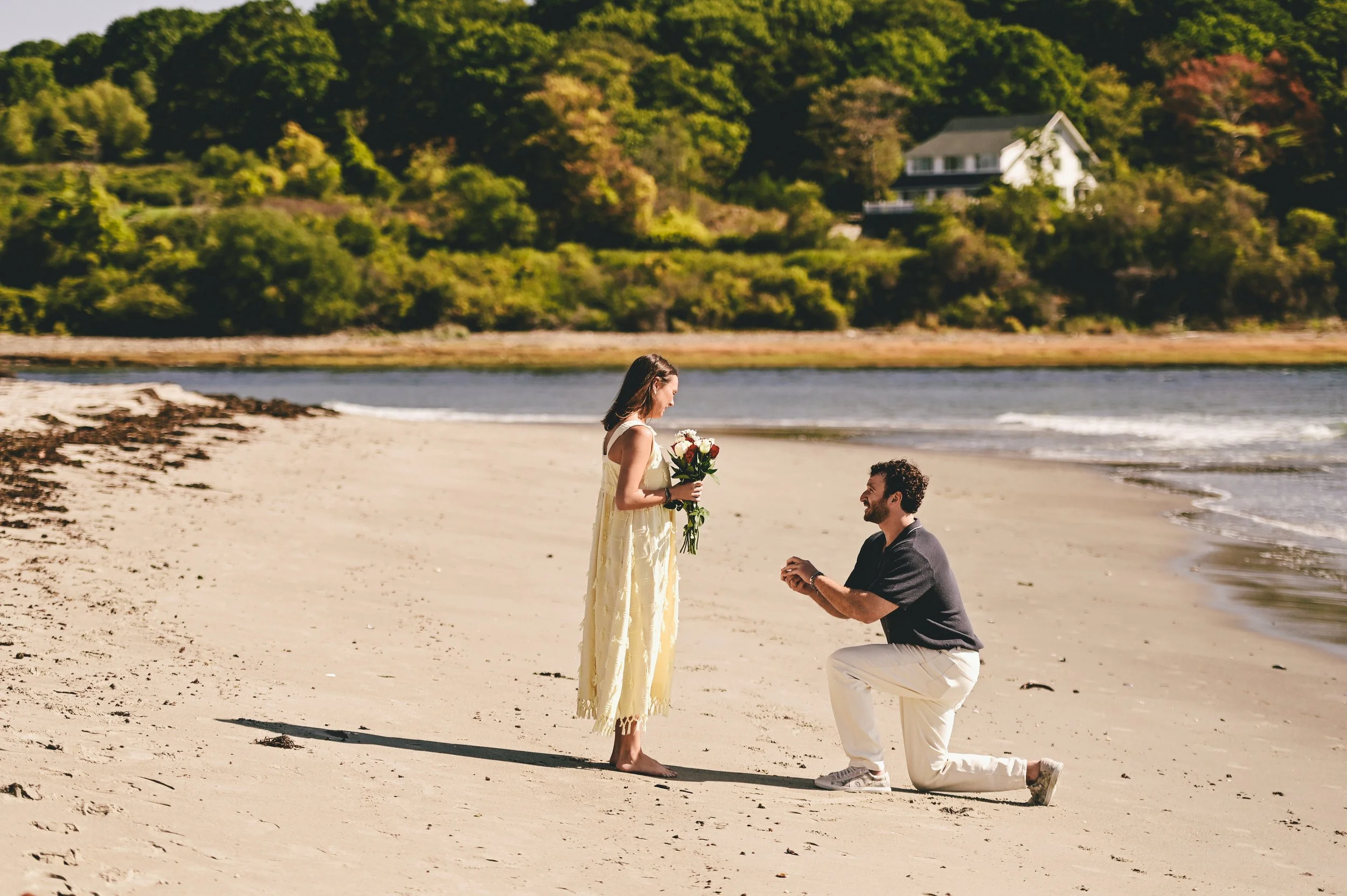 Future groom proposes on sandy coastal beach to future bride standing with bouquet of flowers.