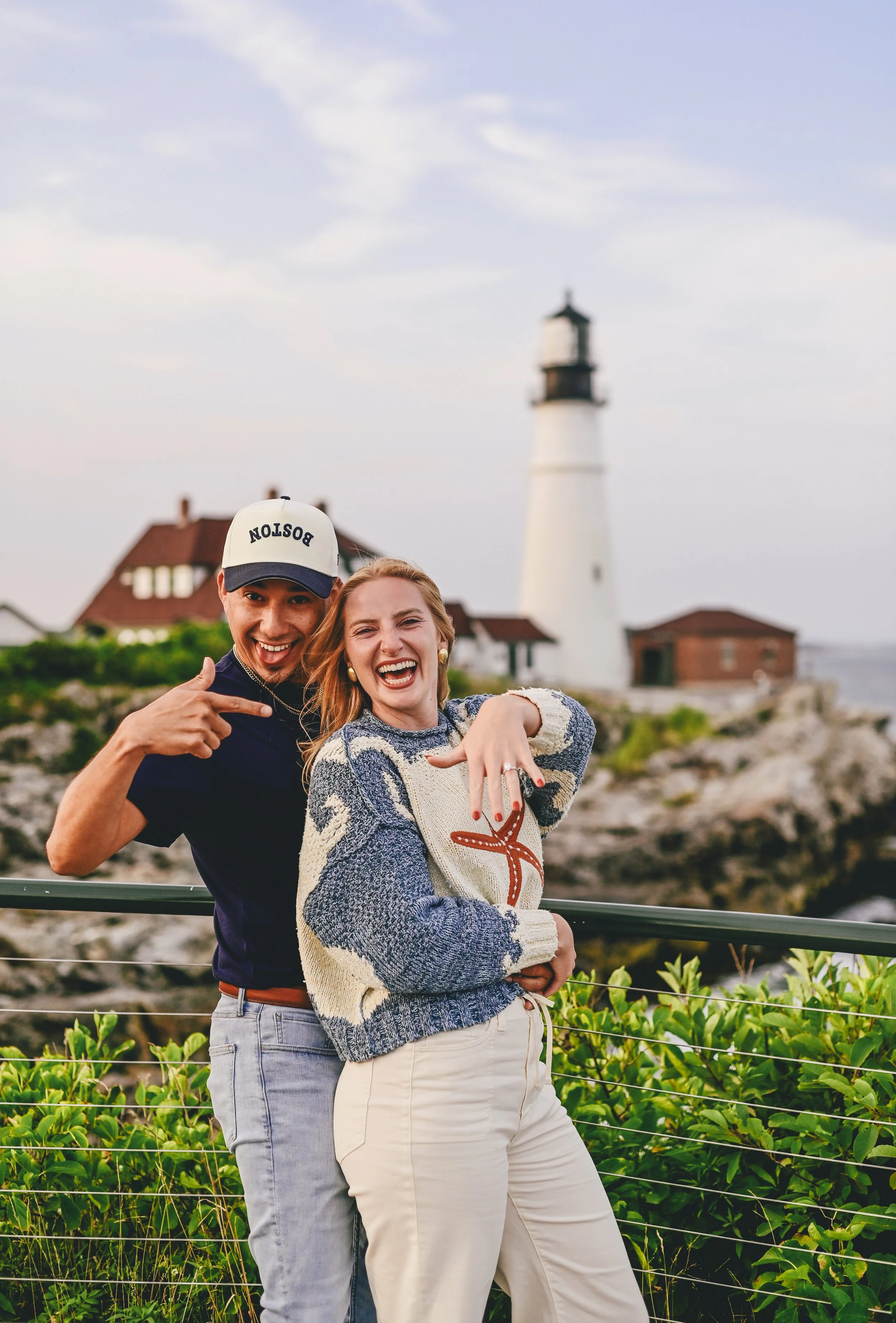 Husband and wife to be celebrate their engagement showing off her ring in front of coastal lighthouse scenery..