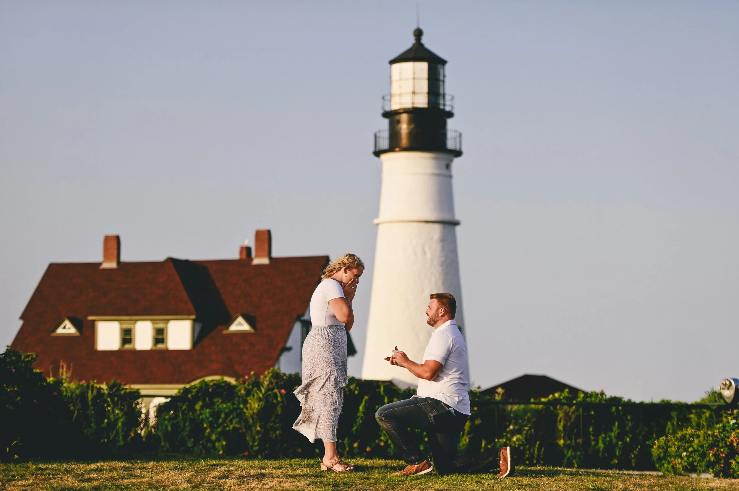 Groom to be proposes on one knee to surprised bride to be in front of Portland headlight lighthouse.