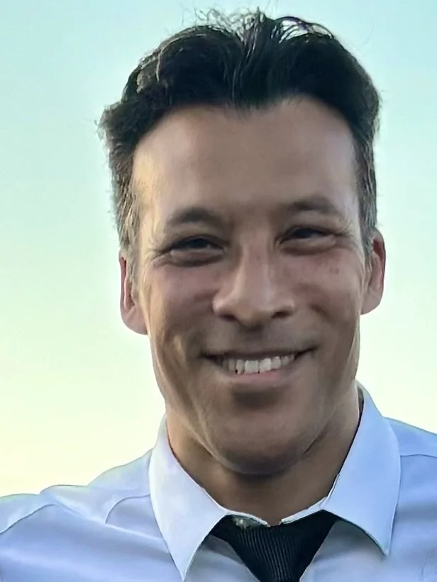 A man smiling outdoors in business attire, wearing a light blue shirt and a dark tie, with a clear sky background.