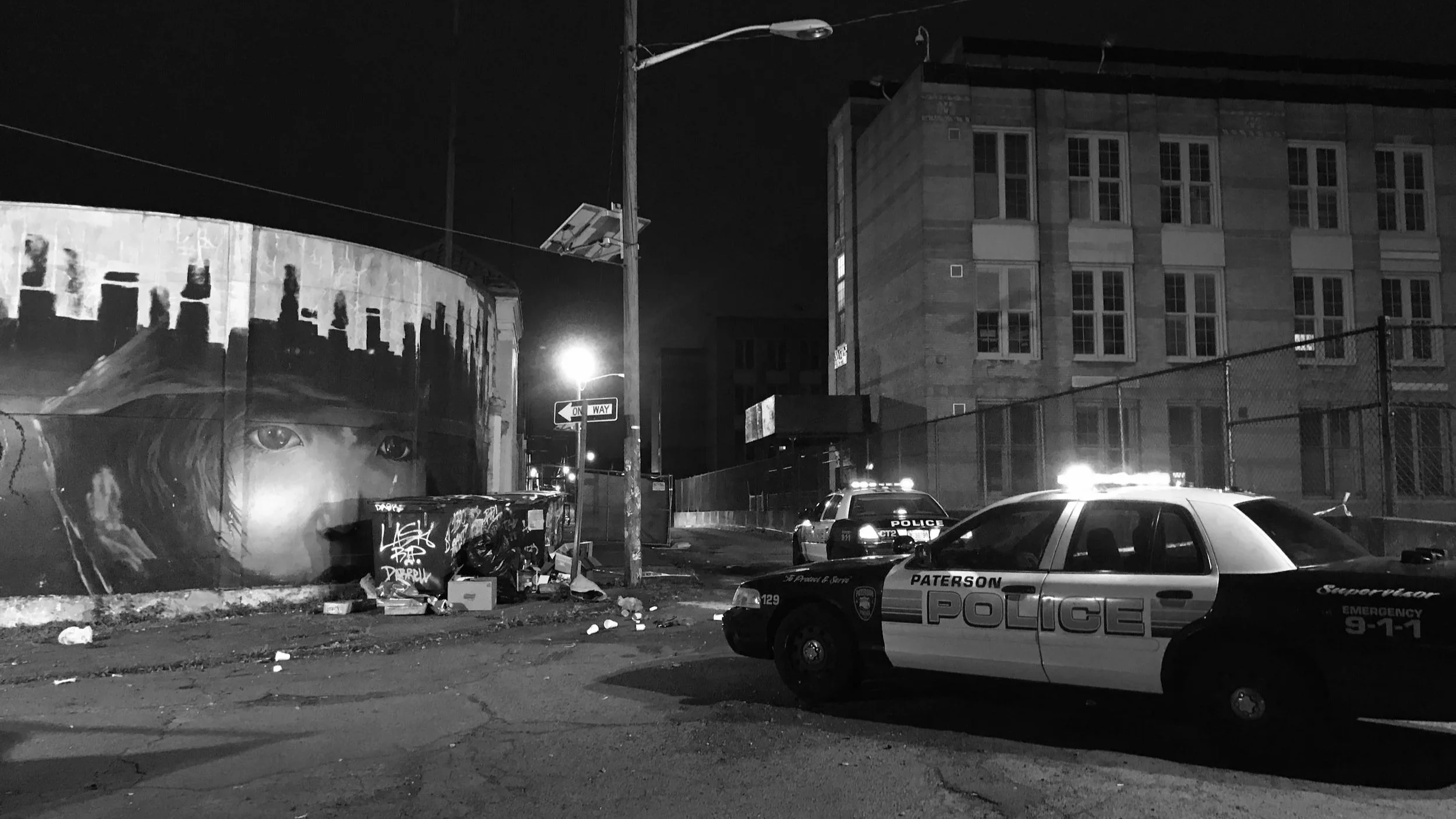 Nighttime scene with two police cars with flashing lights, graffiti mural of a woman's face on a wall, and a building in the background.